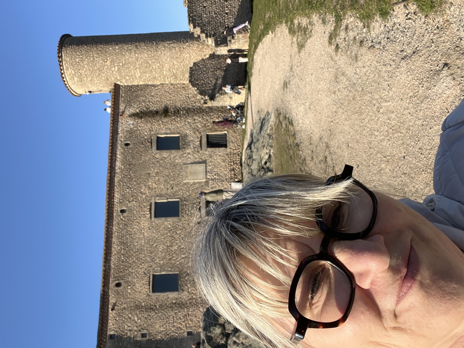 The image depicts an outdoor scene at the Château de Chambles in Chambles, France, under a clear blue sky. In the immediate foreground, centered slightly to the left, is a close-up selfie of a middle-aged individual with light blonde and grey hair, wearing dark-rimmed hexagonal glasses and a light-colored jacket. The individual is looking directly at the camera with a subtle smile.
Behind the foreground subject, the main structure is a large, two-story stone building with a distinctive round tower on its right side. The building features multiple rectangular window openings with dark panes. The roofline has a red tile edge, and two small circular openings are visible higher on the main wall. A light-colored plaque is affixed to the stone wall between the lower-right windows. The lower portion of the tower shows signs of partial ruin.
Several individuals are visible in the mid-ground around the building. Two people are standing on the roof near the tower. On the ground level, to the left of the building, a person is partially visible near a rocky outcrop. Another person in a blue hat is standing near the building's central section. Further right, a person in a dark coat stands near a baby carriage or stroller, with another small group of people nearby. To the far right, a child in purple pants is observed climbing the partially ruined stone wall.
The ground consists of a light-colored, gravel-like path that curves from the lower right towards the center-left of the mid-ground. Areas of green grass and some rocky formations are also present. The scene is illuminated by natural sunlight, casting subtle shadows.