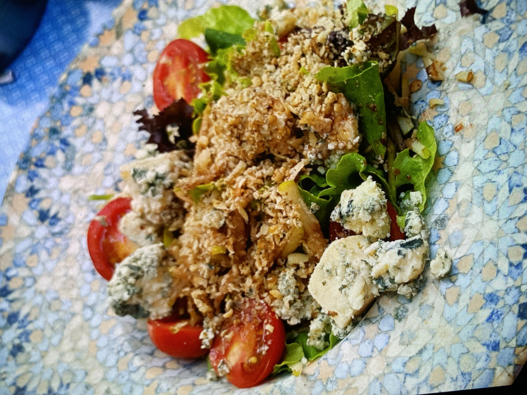 The image displays a vibrant, close-up, top-down view of a fresh salad served on a highly decorative, patterned plate. The salad is composed of various ingredients, including crisp green leafy lettuce, interspersed with some darker, reddish-brown lettuce pieces. Halved red cherry tomatoes are scattered throughout the salad, adding splashes of bright color. Distinct chunks of blue cheese, showing their characteristic creamy white texture with prominent blue-green mold veins, are clearly visible. A generous and chunky topping, appearing to be crumbled nuts (possibly walnuts or pecans) or a savory granola/breadcrumb mixture, is spread over the main body of the salad, providing a significant textural element.
The plate itself is a key visual component, featuring an intricate, mosaic-like pattern in shades of blue, light blue, beige, and cream, which lends an artisanal or Mediterranean aesthetic to the presentation. The image captures the salad ready for consumption, suggesting a mealtime or dining occasion. No specific activity beyond the presentation of food is depicted, nor are any people visible. There is no discernible text within the image. While the time of day and weather conditions cannot be determined, the clear lighting suggests a well-illuminated setting. The context provided indicates the location is Bankya, Bulgaria.