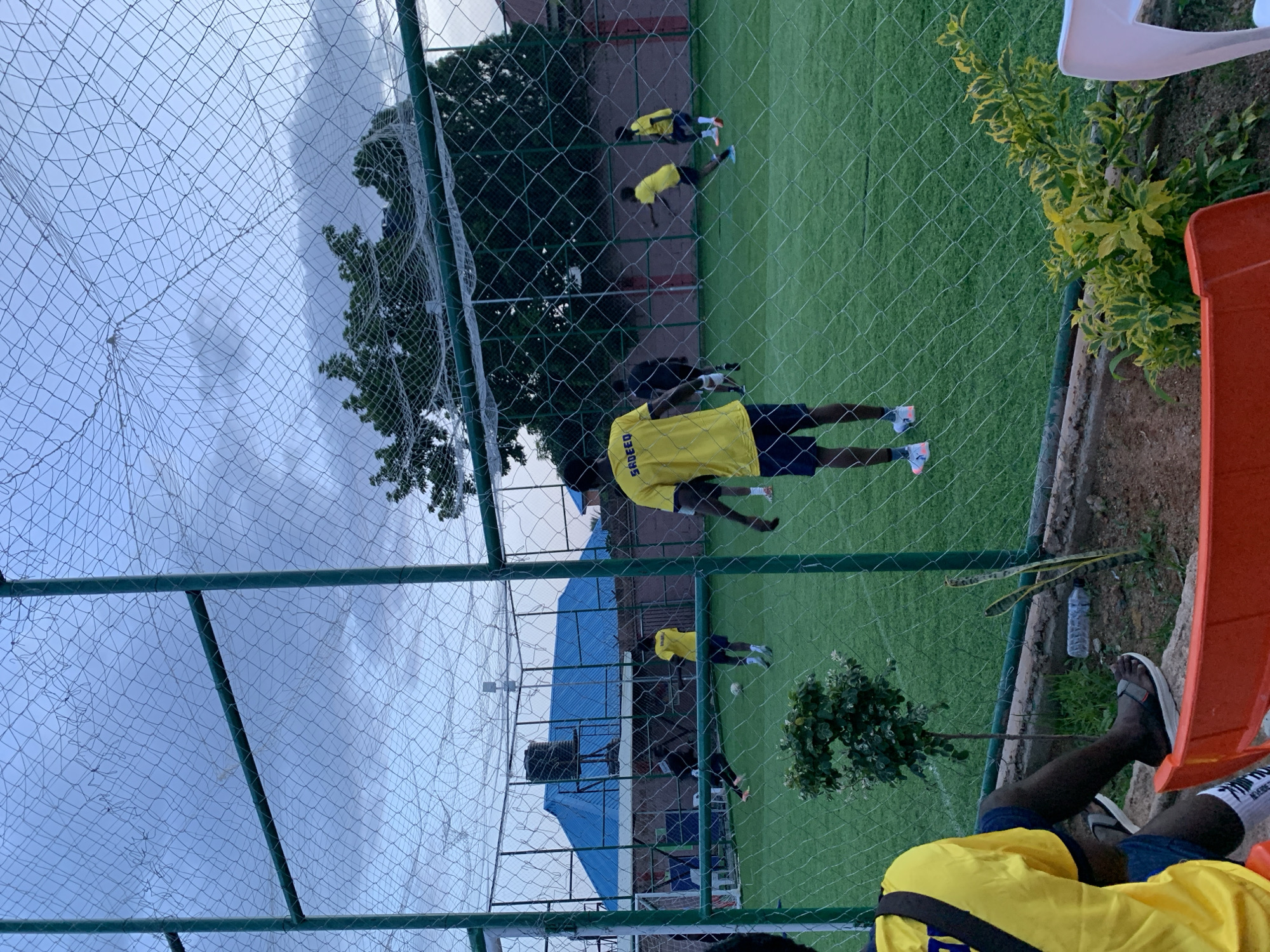 In Gura Top, Nigeria, a group of young men in yellow t-shirts and dark shorts are practicing soccer on an artificial turf field. The scene is enclosed by a green metal fence with netting. In the foreground, a person wearing a yellow t-shirt with "SADEED" written on the back is seen from behind, sitting on an orange chair. In the background, several other individuals are engaged in soccer drills, with one person in the process of kicking a ball. A blue-roofed building with a water tank on top is visible behind the field, along with trees and a cloudy sky. The lighting suggests it is either early morning or late afternoon.