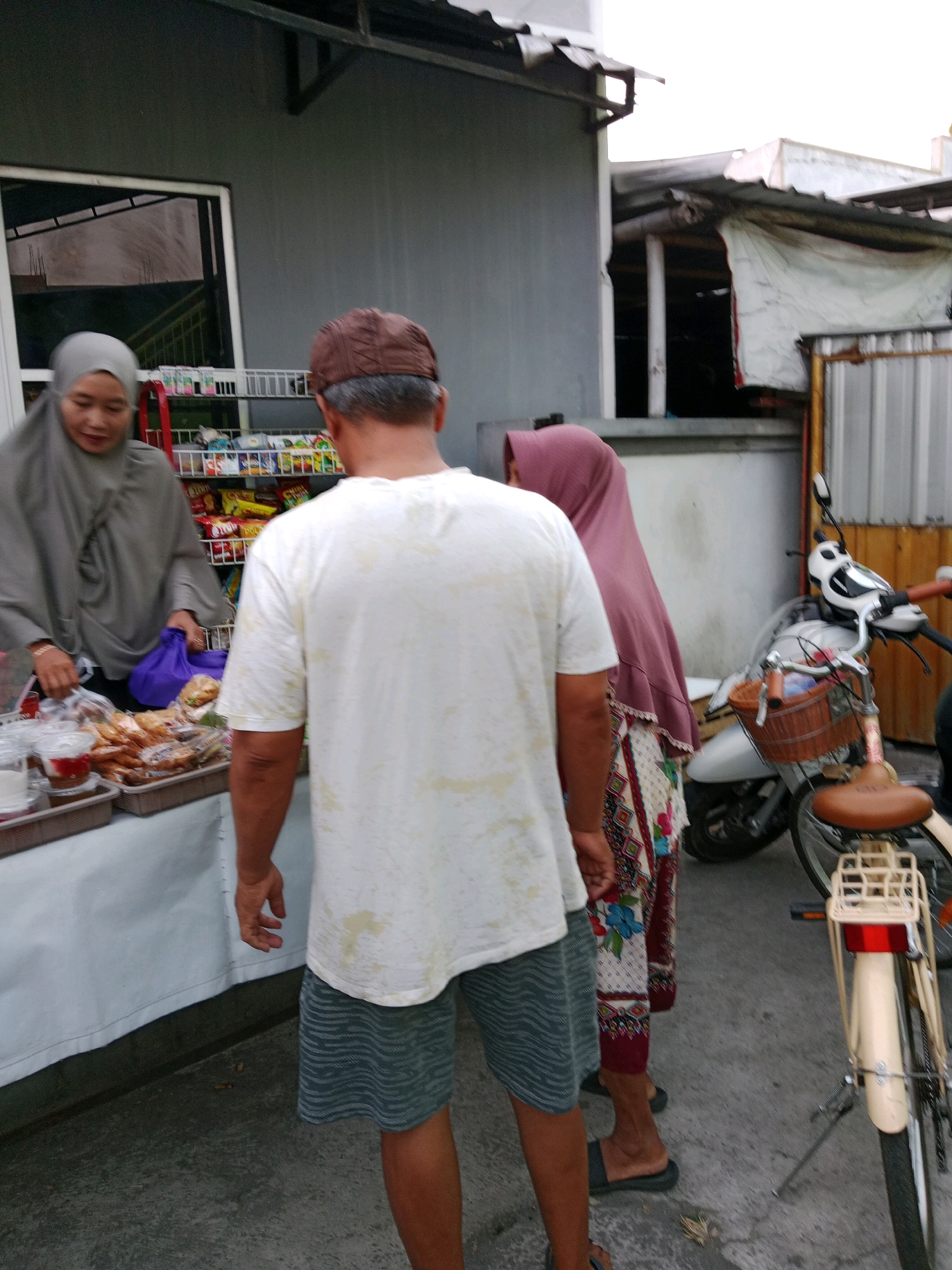 In Waru, Indonesia, an outdoor stall displays various food items. A woman wearing a gray hijab stands behind the stall, attending to a customer, a man wearing a white t-shirt and patterned shorts. The man is facing away from the camera, his back to the viewer, while the woman is looking down, engaged in the transaction. A second woman, partially visible, stands to the right of the man, wearing a pink hijab and a patterned dress. In the background, a white and silver scooter is parked, with a wicker basket on its handlebars. The environment suggests a bustling street market or a roadside vendor.