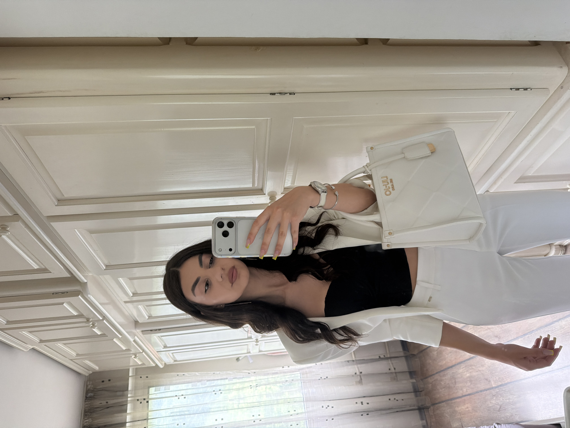 The image depicts a young woman taking a mirror selfie indoors, within what appears to be a residential setting in Gabrovo, Bulgaria. She stands facing forward, holding a silver-colored smartphone with a clear case in her right hand, positioned to reflect her image. Her gaze is directed towards the phone screen, slightly to her left.
The woman has long, wavy, dark brown hair and features a light makeup application with prominent eyebrows and light pink lips. Her manicure features bright yellow nail polish. She is dressed in a formal yet stylish ensemble: a black sweetheart-neckline crop top, a white blazer with three-quarter sleeves, and matching white tailored pants. On her left wrist, she wears a silver-toned wristwatch with a white band and two thin silver bracelets. Her left hand holds a white, structured handbag with a quilted pattern and gold-toned hardware. The handbag has a small tag attached to the handle and a brand name, possibly "LIU JO" or "OL-ULIJ," debossed in gold letters on the front.
The background consists primarily of a wall of light cream or off-white paneled cabinets or wardrobes, extending from the floor to the ceiling. These cabinets feature decorative molding and light-colored handles. Minor scuff marks or streaks are visible on the rightmost cabinet panel near a hinge. To the left of the woman, a window is visible, covered by sheer, light-colored curtains adorned with a subtle pattern of dark square dots. The floor is made of medium-toned wooden planks. The room is brightly lit, likely by natural light emanating from the window.