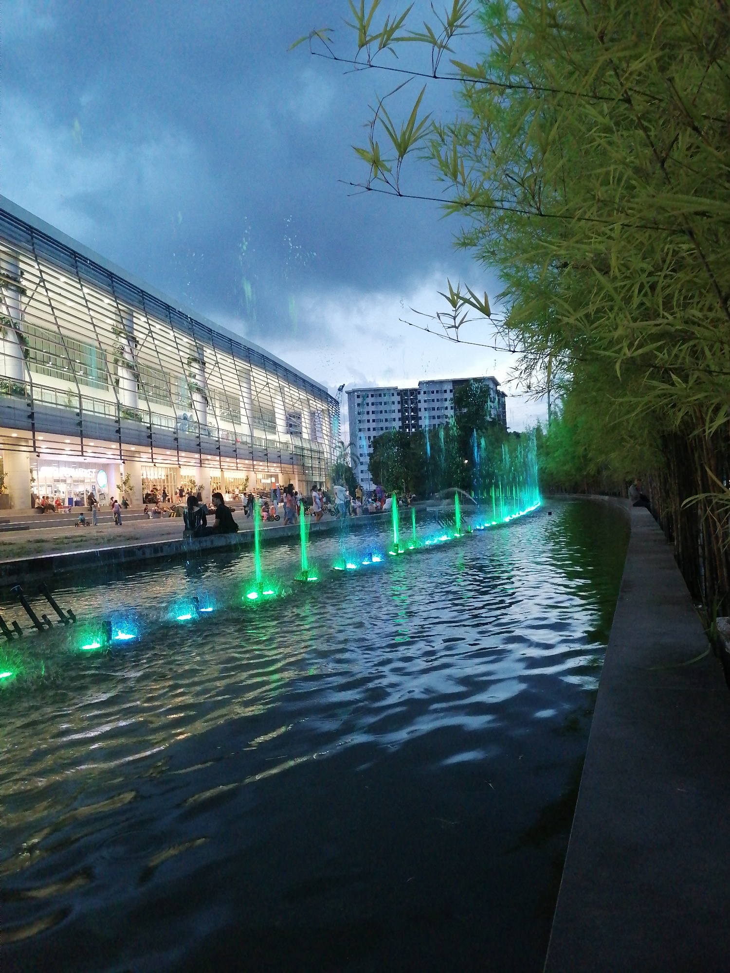 The image captures a twilight scene at SM City Davao's Lanang Park in Davao City, Philippines. A modern, glass-fronted building with prominent horizontal lines, likely a commercial establishment, dominates the left side of the frame. In front of the building, a body of water features a line of illuminated fountains, casting vibrant green and blue light onto the surface. People are visible sitting and walking along the paved walkway adjacent to the water and the building's facade. To the right, a dense grove of bamboo trees creates a natural boundary, with their long, slender leaves illuminated by the ambient light. The sky above is a mix of deep blue and grey clouds, suggesting the late hour. The overall atmosphere is serene and urban, highlighting a recreational space within the city.