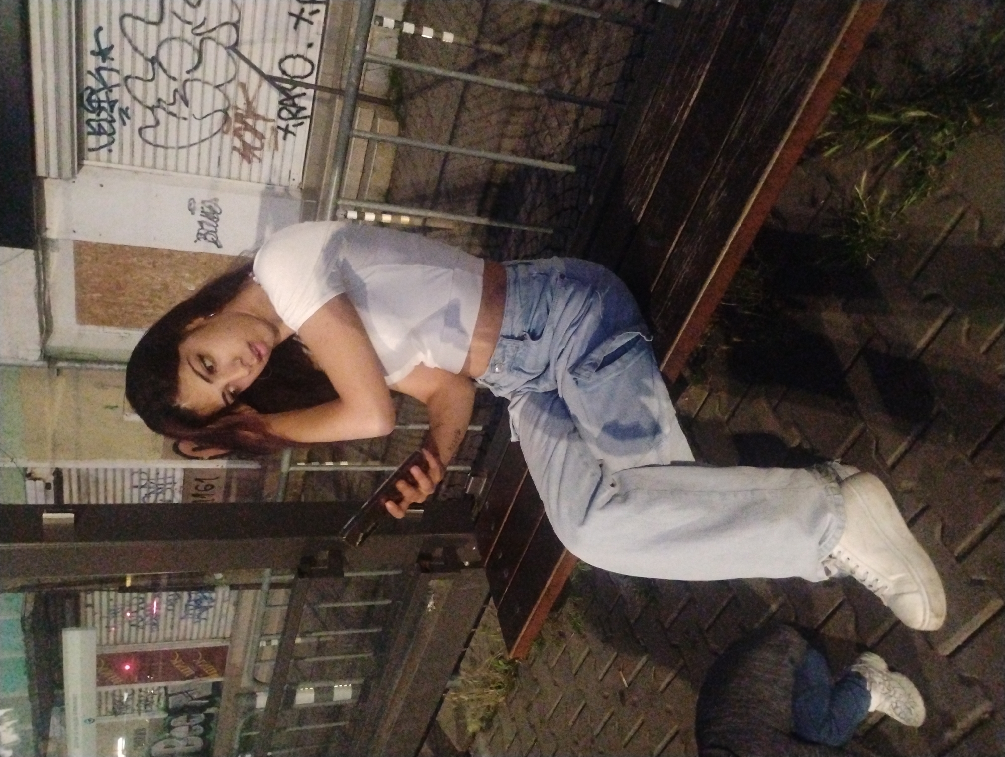 The image depicts an urban scene at night in Sofia, Bulgaria, characterized by dim artificial lighting. A young woman with dark hair and light skin is seated on a dark wooden bench, positioned slightly to the right of the frame's center. She wears a white short-sleeved cropped t-shirt, light blue baggy jeans, and white platform sneakers. Her left hand holds a dark-colored smartphone, while her right hand is raised to the side of her head, appearing to touch or adjust her hair. Her gaze is directed downward or towards the phone. Partially visible in the bottom left corner is a smaller individual, likely a child, wearing a dark top, blue jeans, and white patterned sneakers, crouched on the paved ground. Behind the bench is a dark metal railing. The background consists of buildings with closed metal roller shutters, heavily covered in graffiti. Visible Cyrillic graffiti includes "ШИК" and "TPAIO", along with the number "161". The ground is paved with dark rectangular blocks, with small patches of grass growing near the bench.