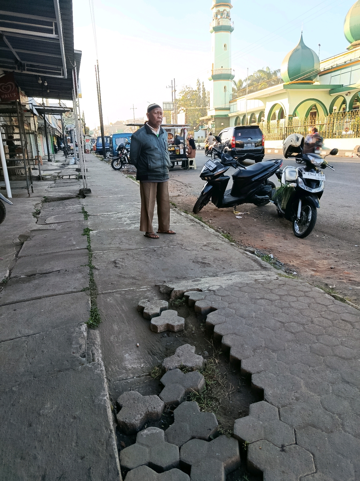 A man stands on a sidewalk next to a street in Bululawang, Indonesia. He is wearing a blue jacket and brown pants. Two motorcycles are parked on the side of the street. A mosque with a green dome and a tall minaret is visible in the background. The sidewalk is made of concrete pavers that are cracked and uneven. There are shops on the left side of the street. The sky is clear and the sun is shining.