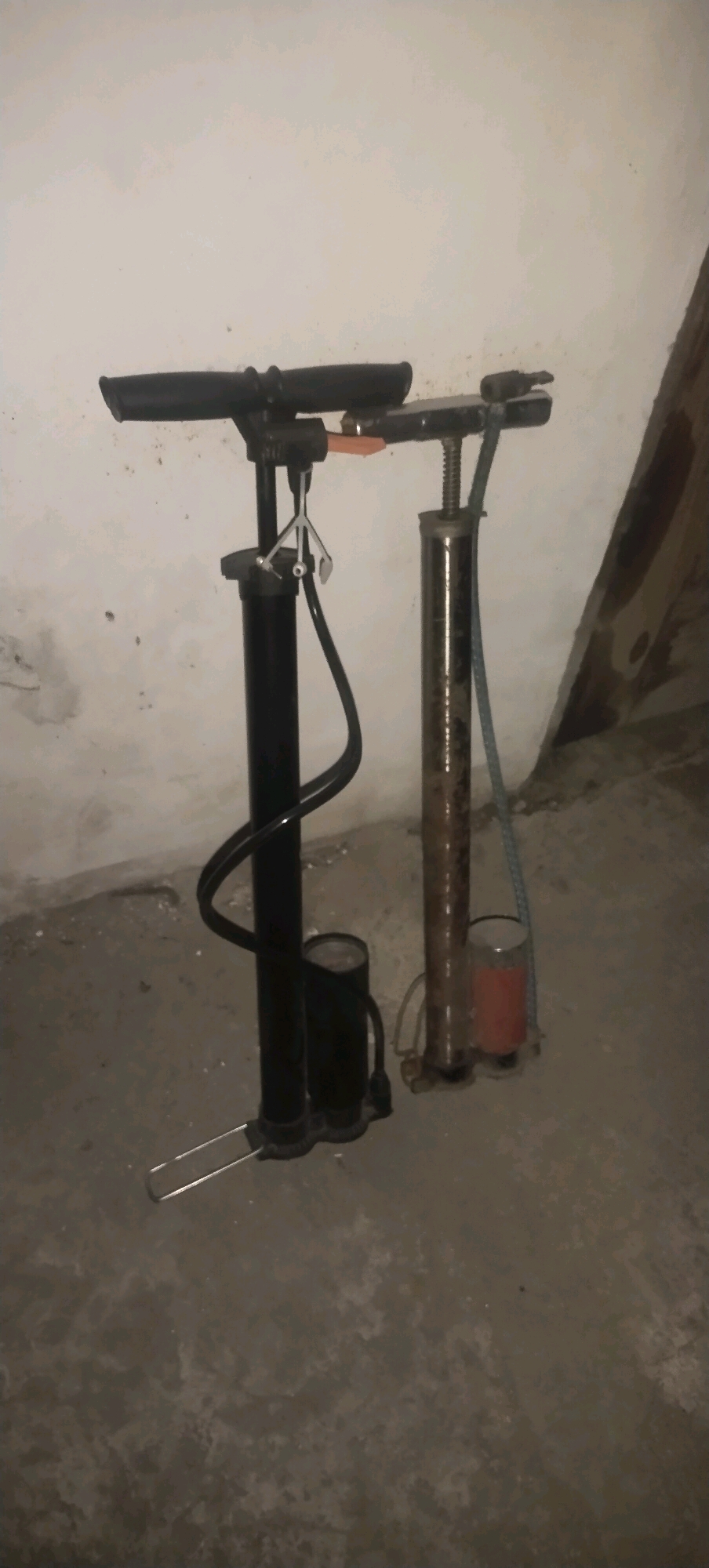 This image shows two bicycle pumps. One pump is black, and the other is silver and rusty. Both pumps are standing on a concrete floor in front of a textured, light-colored wall. The scene appears to be indoors, possibly in a storage area or a garage, as suggested by the wall texture and the unkempt appearance of the floor. The lighting is dim, creating shadows. There are no people visible in the image, nor any actions or interactions taking place. The location of Daleman, Indonesia is not discernible from the visual information provided in the image.