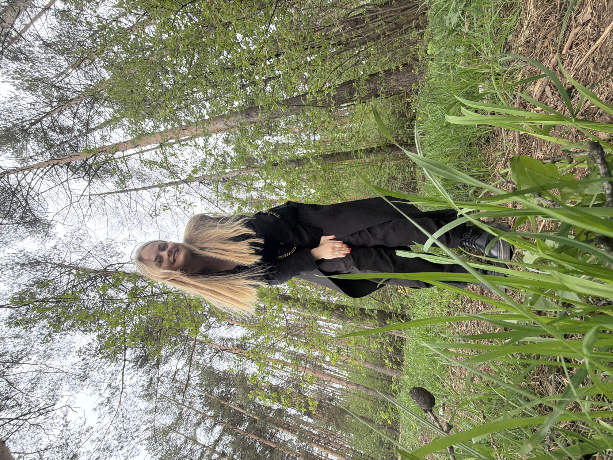 The photograph appears to be taken in a wooded area, possibly within or near Izhevsk, Russia, given the city's location. A woman with long blonde hair is the primary subject. She is wearing a black coat and pants. Her hands are clasped together. She is standing among trees and tall green grass. The background consists of trees with bare branches and some new green leaves, indicating it may be early spring. The sky is overcast.
