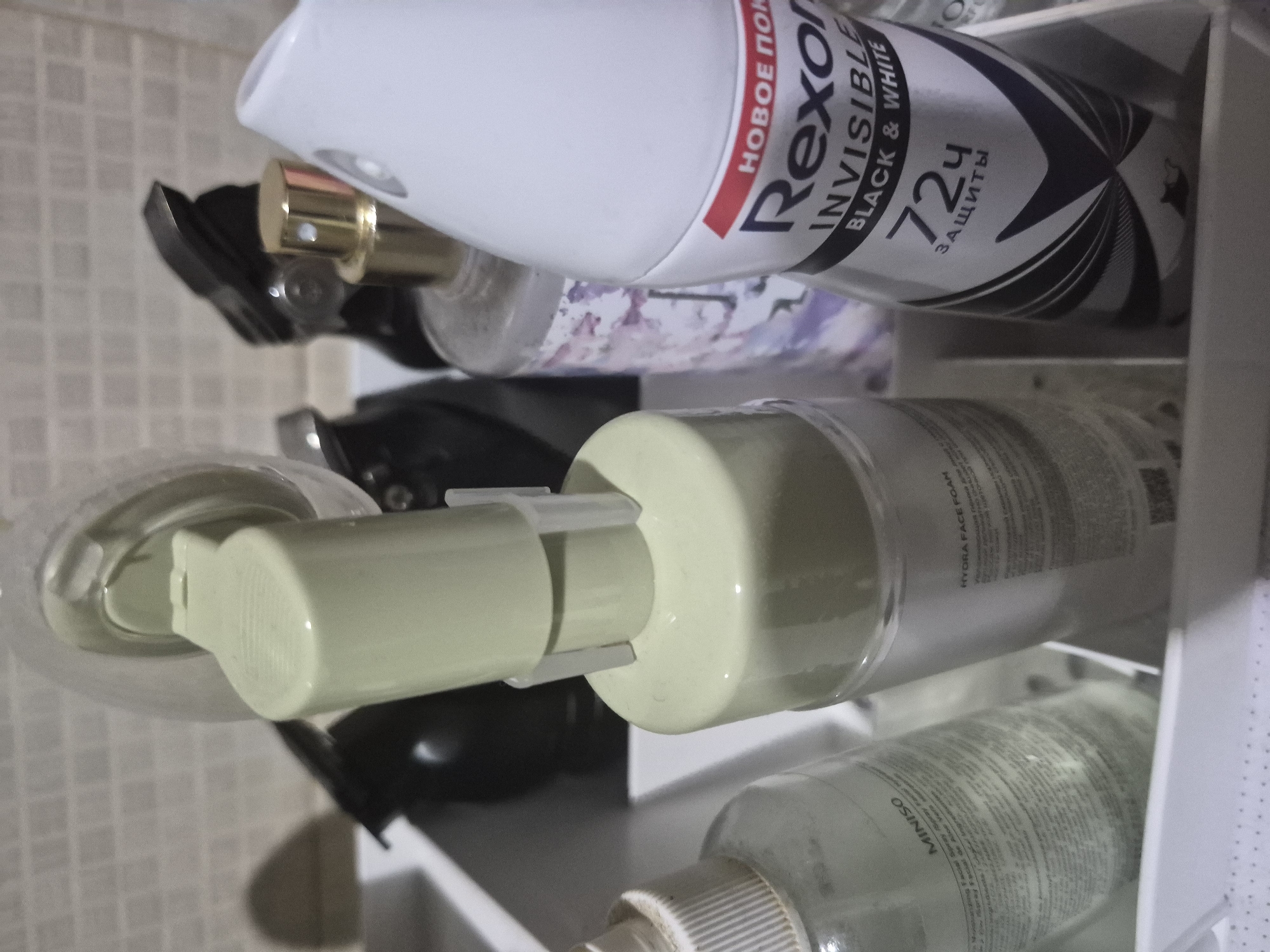This close-up, vertically oriented image captures an assortment of personal care products organized on a white, multi-tiered shelf, set against a background of light-colored, square-tiled wall. The scene suggests a bathroom or vanity area in Yerevan, Armenia.
A prominent white spray can of Rexona deodorant occupies the upper part of the shelf. Its label clearly displays "НОВОЕ ПОК" (New cover/coating), "Rexona", "INVISIBLE BLACK & WHITE", and "72Ч ЗАЩИТЫ" (72 hours protection), indicating a 72-hour invisible antiperspirant. Below this, a clear bottle with a gold-colored spray or pump mechanism is partially visible. Further down, a light green pump bottle, likely a facial cleanser, shows the partial text "HYDRA FACE FOAM". On the lowest visible shelf, a white bottle with a white cap is identifiable by the "MINISO" brand name printed on its label. Other bottles are blurred in the foreground and background.
No individuals are depicted, and no specific activity is taking place; the image focuses solely on the static arrangement of these everyday toiletries. The lighting is soft, creating some shadows among the items.