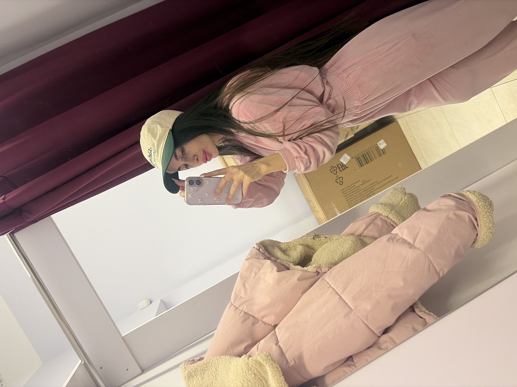 A female individual is depicted taking a mirror selfie inside what appears to be a retail store changing room in Saint Petersburg, Russia. The subject has long, dark hair and is wearing a light pink velvet tracksuit with a beige baseball cap featuring a dark green brim. She holds a light purple smartphone, adorned with flower-shaped embellishments, in her right hand, extending her arm to capture her reflection. Her lips are pursed in a pose. To the left, a light pink quilted puffer jacket with a cream-colored fleece lining hangs on a rail, partially visible in the mirror. A large, unsealed cardboard box displaying a barcode and Cyrillic text (e.g., "ЕАЭС," "РУС") rests on the floor in the reflected background. Burgundy-colored curtains are present on the right side of the frame. The changing room features light-colored walls, a tiled floor, and a metal hanging rail at the top. A small white device, possibly a sensor, is affixed to the wall in the upper reflected area.