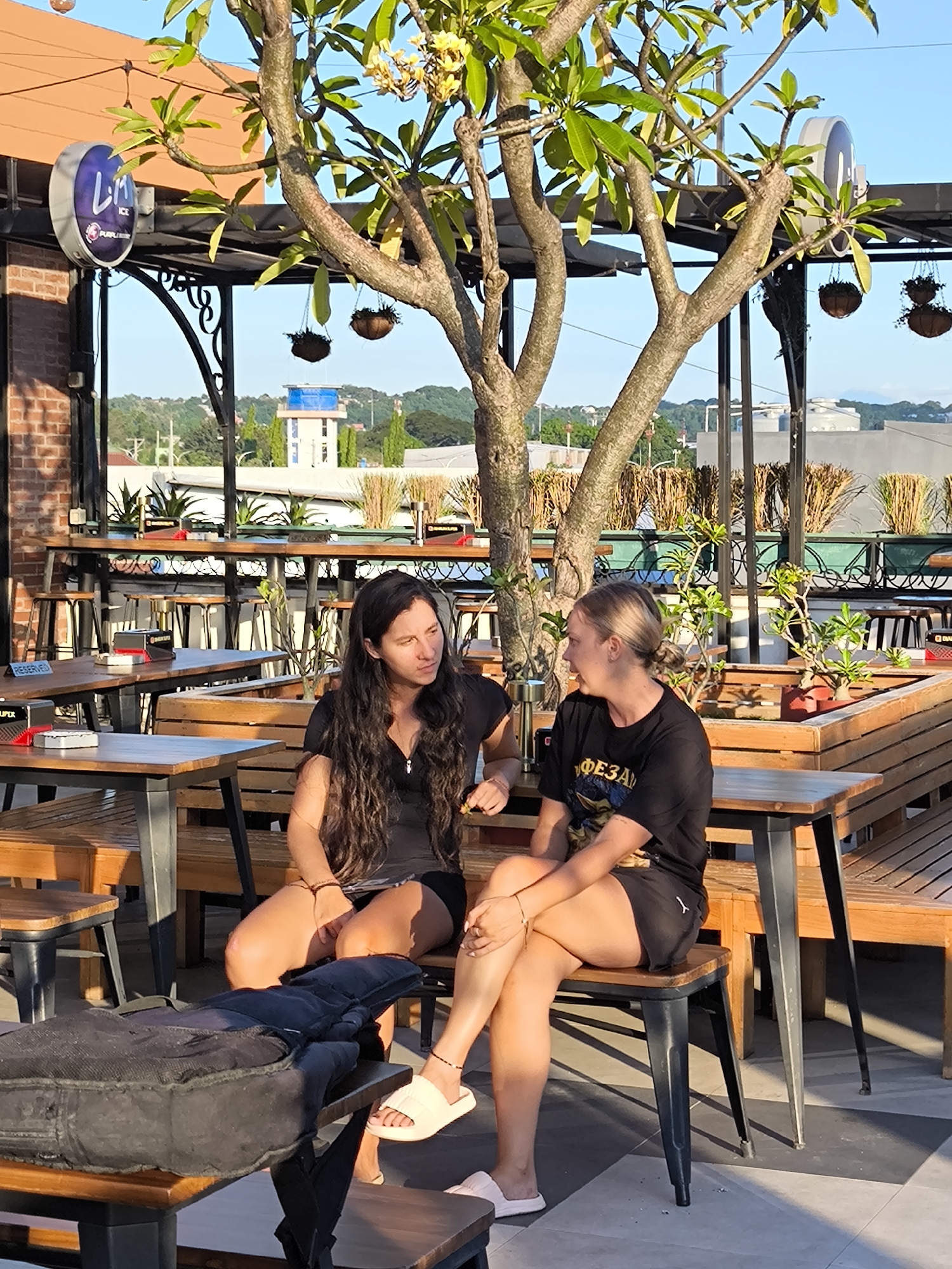 Two women sit at a wooden table on a rooftop patio in Sumbawa Besar, Indonesia. The woman on the left has long, dark hair and wears a black dress. The woman on the right has blonde hair in a bun and wears a black t-shirt and shorts. They both have bracelets and are facing each other in conversation. A black bag sits on a small table next to them. The patio has wooden benches and tables, with various plants and decorative items around. In the background, buildings and trees are visible. The patio is partially covered by a metal awning with a tree growing through the center. A sign is visible on the left that reads "Purple Ice".