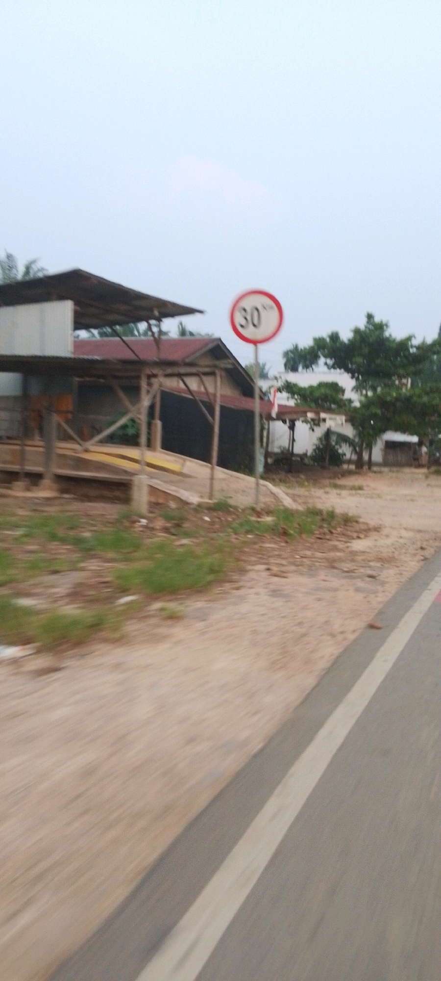 This is a vertical, slightly blurry image taken from a moving vehicle, likely a motorcycle or car, looking towards a rural or semi-rural area in Lenggadai Hulu, Indonesia. In the foreground, a paved road with a white line marking the lane is visible, curving slightly to the right and extending into the distance. The road appears to be made of asphalt and is in reasonable condition.
To the left of the road, a dirt and gravel area is visible, with patches of dry grass and some scattered debris. Beyond this, there is a simple, open-sided structure with a corrugated metal roof and wooden supports, possibly a shelter or a small workshop. In front of this structure, a ramp-like feature made of wood and yellow material is present. Further behind this structure, a house with a red tiled roof and dark walls is partially visible, along with some trees.
A prominent object in the scene is a speed limit sign, circular with a white background and a red border, displaying "30" in black text with "KM/H" below it. The sign is mounted on a metal pole. Several trees, some with dense foliage, are scattered throughout the background. The sky is overcast and pale. The overall impression is of a quiet, possibly somewhat neglected, roadside environment.