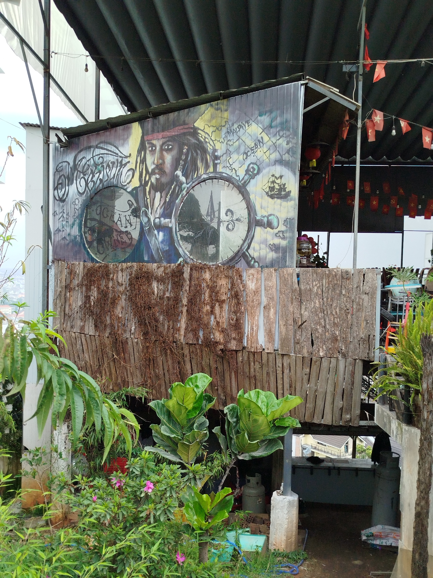 This image captures a rustic, multi-level structure, likely an informal cafe or shop, nestled amidst lush greenery in Da Lat, Vietnam. The upper part of the building features a prominent and colorful mural painted on corrugated metal siding.
The central subject of the mural is Captain Jack Sparrow from *Pirates of the Caribbean*, depicted with his characteristic dreadlocks, bandana, and goatee. He is positioned between two large circular elements that resemble compasses or gears. On the left circular element, the word "OCEAN" is clearly visible, along with a partial "UGAN". On the right, a stylized map shows text including "MALDIVES" and "OCEAN", accompanied by a drawing of a sailing ship. Above Jack Sparrow's head, there is intricate, stylized graffiti text. The overall mural design evokes a maritime and adventurous theme.
Below the painted metal, the structure is built with textured, weathered wooden planks and strips of bark, giving it a raw and natural appearance. The area around the building is verdant, with various plants, including large-leafed foliage, ferns, and bushes bearing pink flowers (likely azaleas), creating a garden-like atmosphere.
In the background, under a metal roof, numerous red flags with yellow stars, characteristic of the Vietnamese national flag, are strung along lines. Some flags also display the yellow hammer and sickle emblem. The scene is illuminated by daylight, suggesting a bright but possibly overcast day. Visible at the base of the structure are propane tanks, indicating practical utility. Distant residential buildings can be seen through openings, suggesting an elevated or hillside location for the structure.