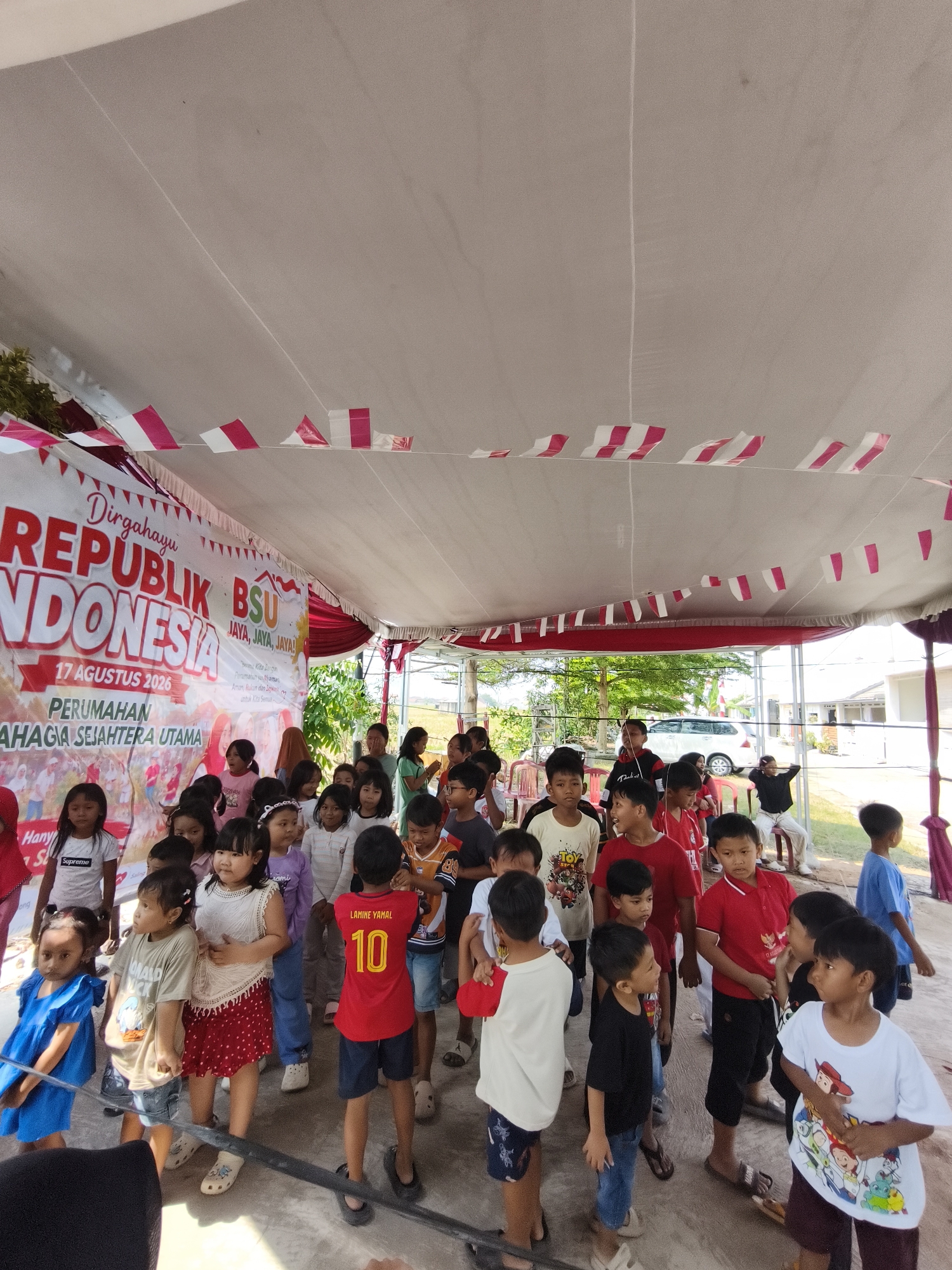 A group of children are gathered under a white tent decorated with red and white Indonesian flags. A banner in the background reads "Dirgahayu REPUBLIK INDONESIA 17 AGUSTUS 2026." The children appear to be participating in an outdoor event, possibly a celebration or gathering. Some children are looking at the camera, while others are engaged in conversation or activity. The environment suggests a warm, sunny day. The location is identified as Bandar Lampung, Indonesia, a city known for its vibrant culture and Independence Day celebrations.