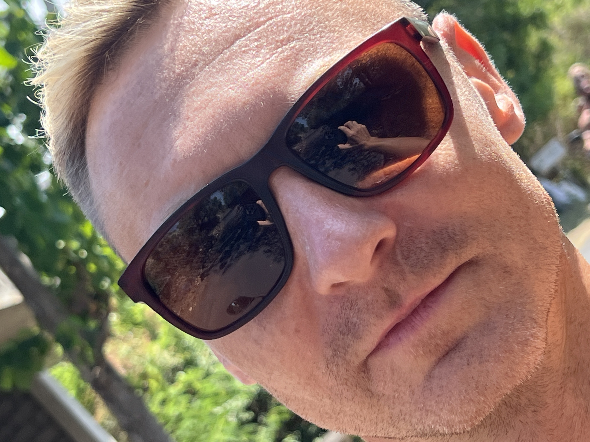 A close-up, over-the-shoulder shot captures a person wearing dark-rimmed sunglasses. The sunglasses reflect an outdoor scene with greenery and what appears to be a hand holding a phone, suggesting the photo is a selfie taken in a location with natural surroundings. The individual's skin is visible, with some stubble on the chin and sideburns, and their hair is light brown and styled upward. The background is softly blurred, showing out-of-focus trees and foliage, indicative of an outdoor setting. The image does not provide any specific information about the location in Abang, Indonesia.