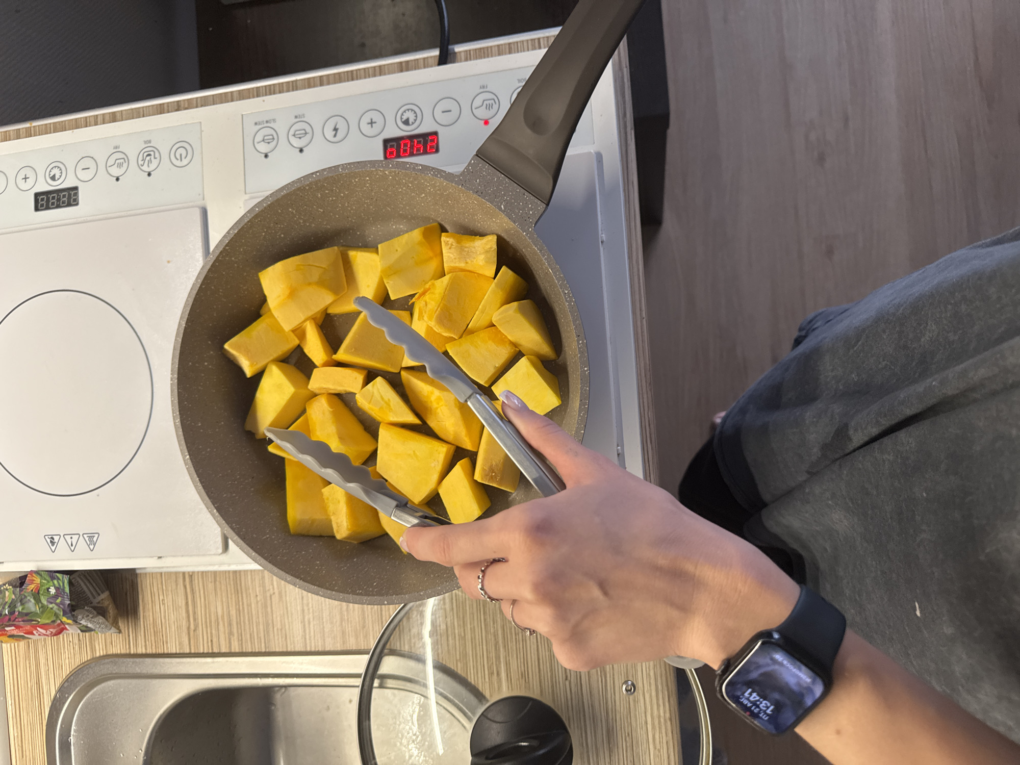 A person's hand, wearing two rings and an Apple Watch displaying "ПТ, 21 авг 13:41", is using tongs to arrange cut pieces of pumpkin in a gray frying pan. The pan is placed on a white induction cooktop with a digital display showing "0:00". To the left of the cooktop is a stainless steel sink with a glass lid beside it. In the background, a bag of what appears to be spices or dried food items is visible. The cooktop is situated on a wooden countertop. The overall environment suggests a kitchen setting. The image does not provide any information about the city of Perm, Russia.