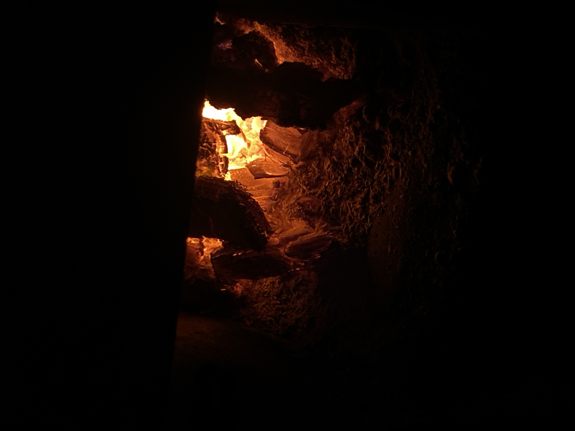 The image shows the inside of a fireplace, with glowing embers and burning logs visible. The fire is intense, with flames casting a warm orange glow on the surrounding wood and ash. The scene is dark, with the only light source being the fire itself. The fireplace appears to be made of brick or stone. The image does not provide any information about the city of Nyagan, Russia, or any people or actions taking place. It is a still shot of an active fire within a fireplace.