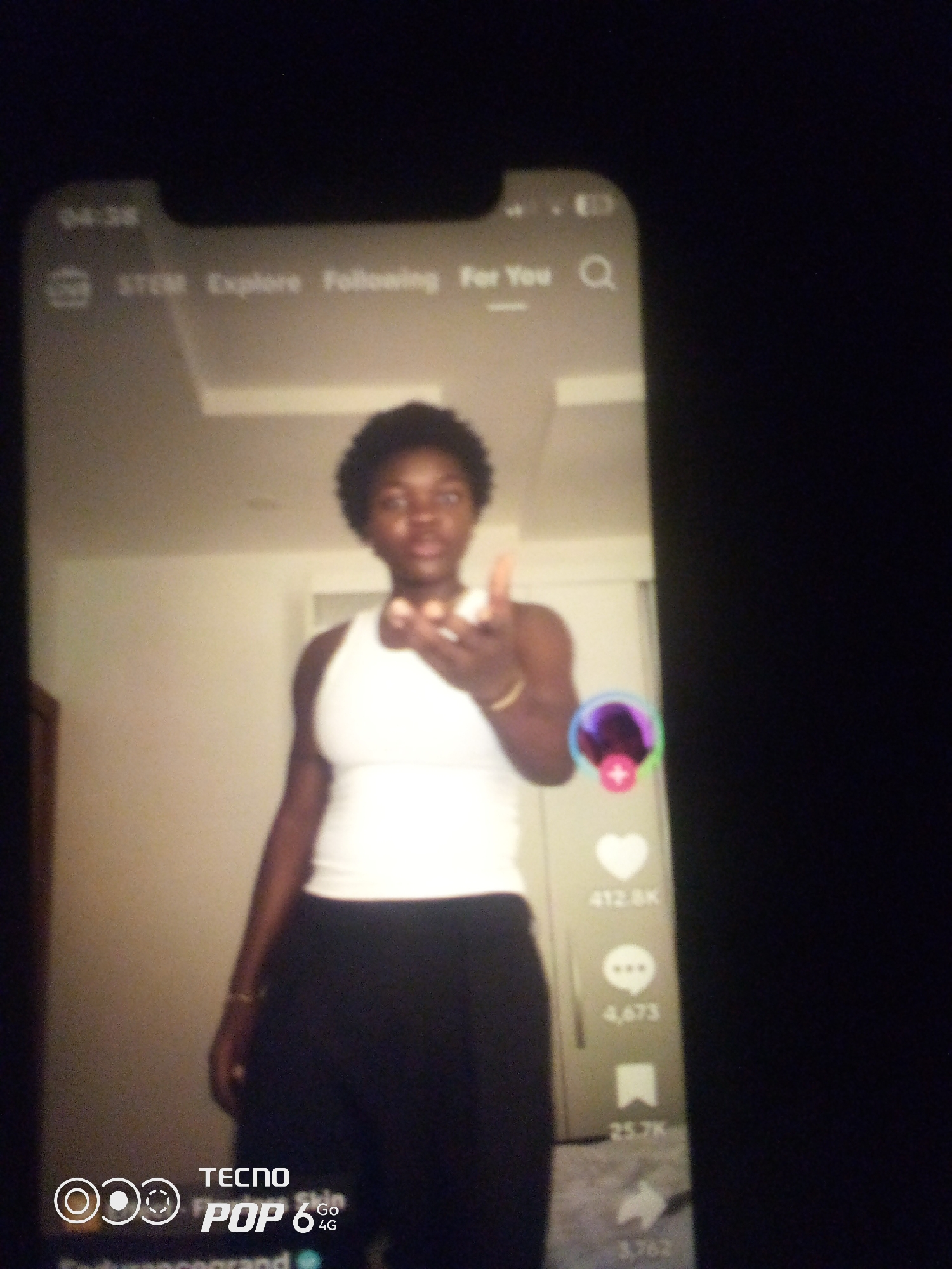 This is a medium shot of a young Black person, with short, dark, curly hair, wearing a white tank top and dark pants, standing in what appears to be a room. The person is facing the camera and extending their right hand towards it. Their expression is neutral, and they appear to be posing for a photo or video. The background is softly lit, with a ceiling that has square panels, suggesting an indoor setting.
The image is a screenshot of a mobile phone screen displaying a social media application, likely TikTok given the layout of icons and text on the right side. Visible text includes "STEM," "Explore," "Following," and "For You" at the top, along with a search icon. On the right side of the screen, there are engagement metrics: a heart icon with "412.8K" (likely likes), a speech bubble icon with "4,673" (likely comments), and a bookmark icon with "25.7K" (likely saves). At the bottom of the screen, "TECNO POP 6 Go 4G" is displayed, indicating the phone model. The time on the phone screen reads "04:38."
The overall mood is casual and personal, as if the person is sharing content from their device. The lighting is somewhat dim, suggesting it might be evening or indoors with artificial light. There are no specific weather cues. The location context "Howayi, Nigeria" is provided externally and does not have direct visual cues within the image itself.