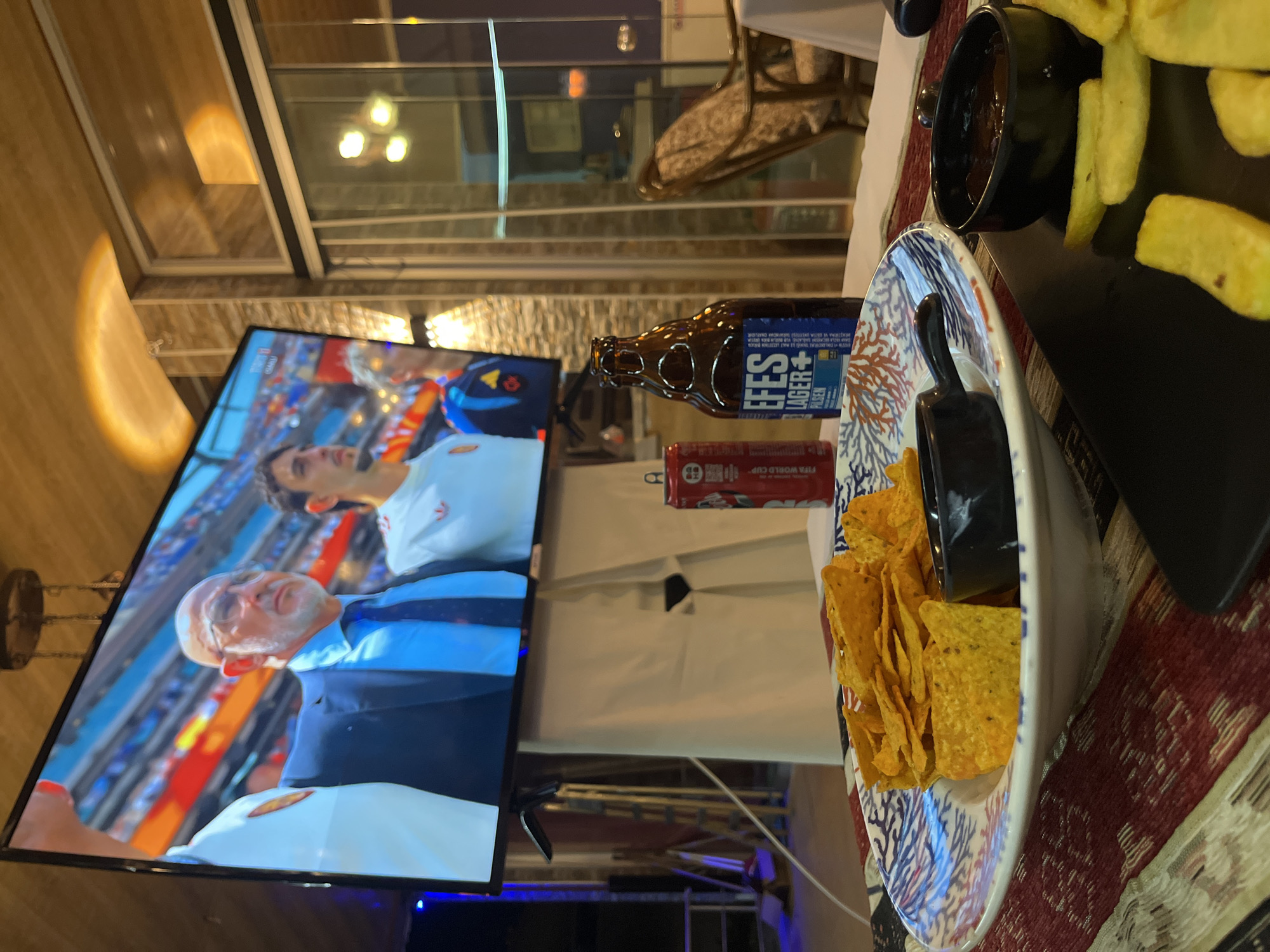 The image shows a low-angle, eye-level shot of a television displaying a soccer match, with a plate of nachos and a bottle of Efes beer in the foreground. The television screen shows two men, one with a bald head and glasses, wearing a dark suit and tie, and another younger man in a white soccer jersey. The background of the television screen depicts a stadium crowd.
In front of the television, a white bowl with a blue and red coral pattern contains a pile of tortilla chips (nachos). Adjacent to the nachos is a small black bowl with a dark dipping sauce. To the right of the nachos is a dark brown glass bottle of Efes Pilsen Lager beer with a blue and white label. Behind the beer bottle is a red can with white lettering.
The table in the foreground is covered with a runner featuring a striped pattern of red, white, and beige. The floor in the foreground has a similar striped pattern.
The environment appears to be an outdoor or semi-outdoor seating area, possibly a restaurant or bar. In the background, there are glass doors and windows, suggesting an enclosed space. A wooden ceiling with exposed beams and lighting fixtures is visible. To the right, a wicker chair with a patterned cushion is partially visible. The overall lighting is dim, with warm yellow light reflecting off the ceiling and illuminating the scene. The presence of the Efes beer, a popular Turkish brand, suggests the location is likely in Turkey. Considering the context of an establishment serving Efes beer and displaying a soccer match, it is plausible this scene is at a bar or restaurant in Alanya, Turkey.