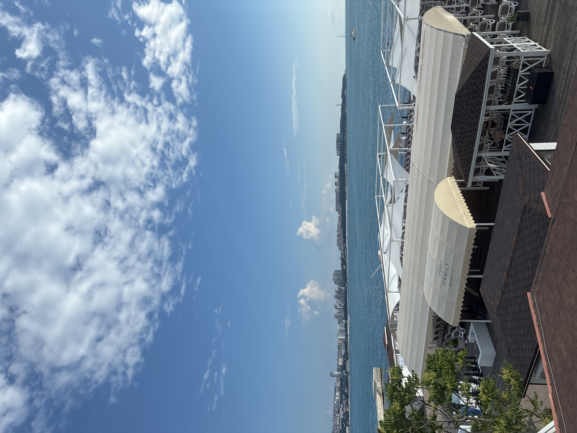 This is an outdoor, eye-level, vertical, daytime photograph taken from a rooftop overlooking a resort area near the sea. The scene is a resort with white tents providing shade over lounge chairs. In the foreground, there are rooftop structures with brown shingles. To the left, a tree with lush green foliage grows, and beyond that, a giant chess set is visible on a wooden deck. A man sits on a white sofa. In the middle ground, the sea is calm and blue, with a distant city skyline on the horizon. The sky is a vibrant blue with scattered white clouds. In the middle ground, to the right of the city skyline, a sailboat is visible on the water. The sign on one of the awnings reads "alean FAMILY BIARRITZ". This resort is likely located on the Gelendzhik Bay, a popular resort city in Krasnodar Krai, Russia.