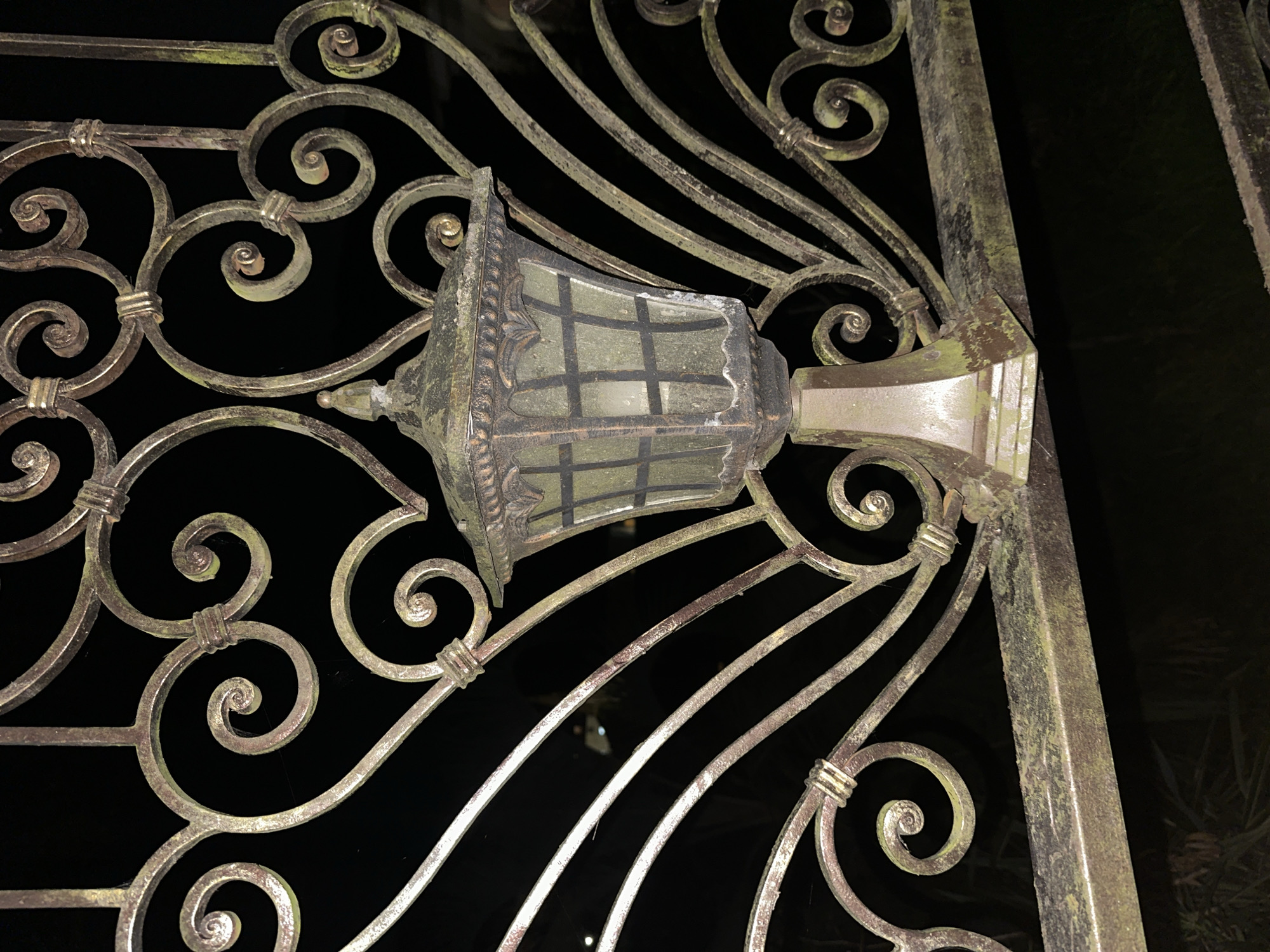 A close-up, low-angle shot captures an antique, ornate metal gate in Sochi, Russia. The gate, with its intricate scrollwork and heart-shaped designs, appears weathered and possibly covered in a patina of moss or dirt, giving it a rustic, aged look. Mounted on the gate is a vintage-style lantern with a multi-paneled glass enclosure, its light faintly visible within. The background is dark, suggesting the photograph was taken at night or in a dimly lit area, with blurred hints of foliage and distant lights visible through the gate's openings. The focus is on the texture and detail of the gate and lantern, highlighting their decorative elements and signs of age.