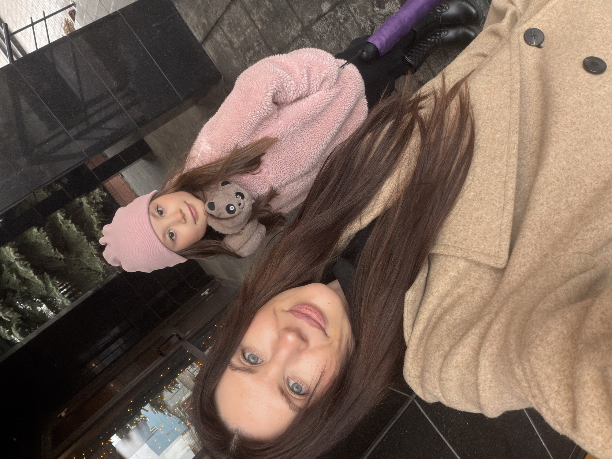 The image shows a selfie taken by a woman with long brown hair and blue eyes. Next to her is a young girl wearing a pink hat and a pink furry jacket, holding a stuffed panda toy. The background appears to be an urban environment, possibly in Perm, Russia, based on the architectural style and general atmosphere. Reflections in a dark, tiled surface suggest a building with lit windows and possibly holiday decorations. The overall scene is casual and likely depicts a mother and daughter posing for a photo outdoors in an urban setting. The angle suggests the subjects are lying down. There is also a purple leg cover and black shoes in the image.