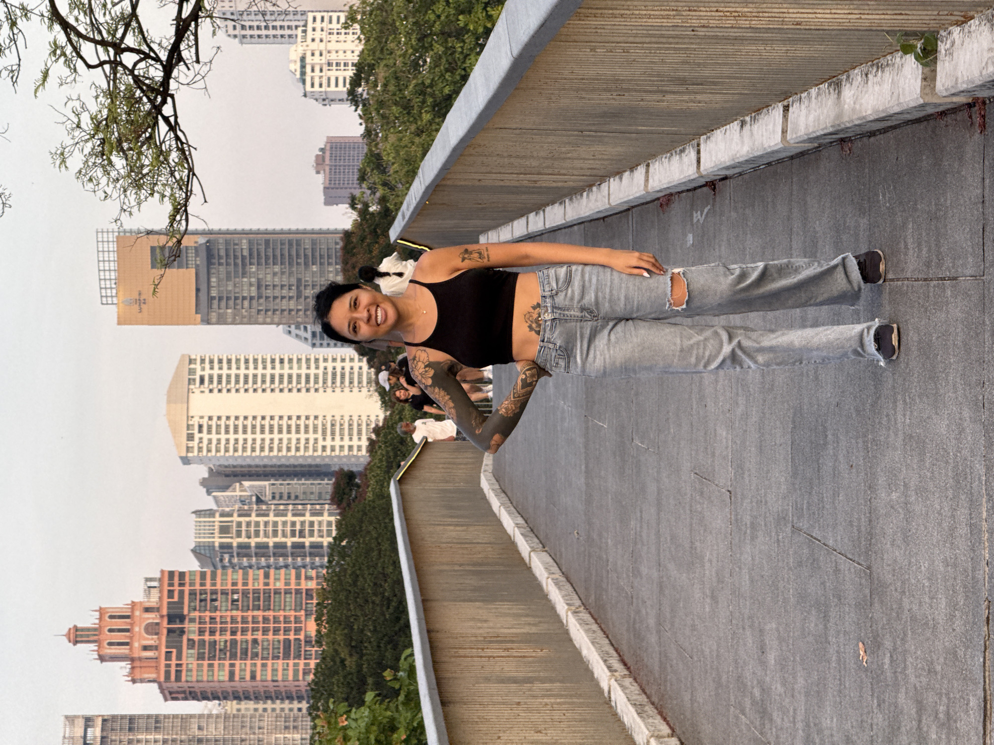 A young woman with dark hair pulled back, wearing a black tank top and ripped jeans, stands on a concrete pedestrian walkway in Bangkok, Thailand. She is smiling and facing the viewer. The woman has tattoos on her arms, torso, and upper arm. The walkway is flanked by a low concrete wall. In the background are numerous high-rise buildings, trees, and some other pedestrians. The sky is overcast.