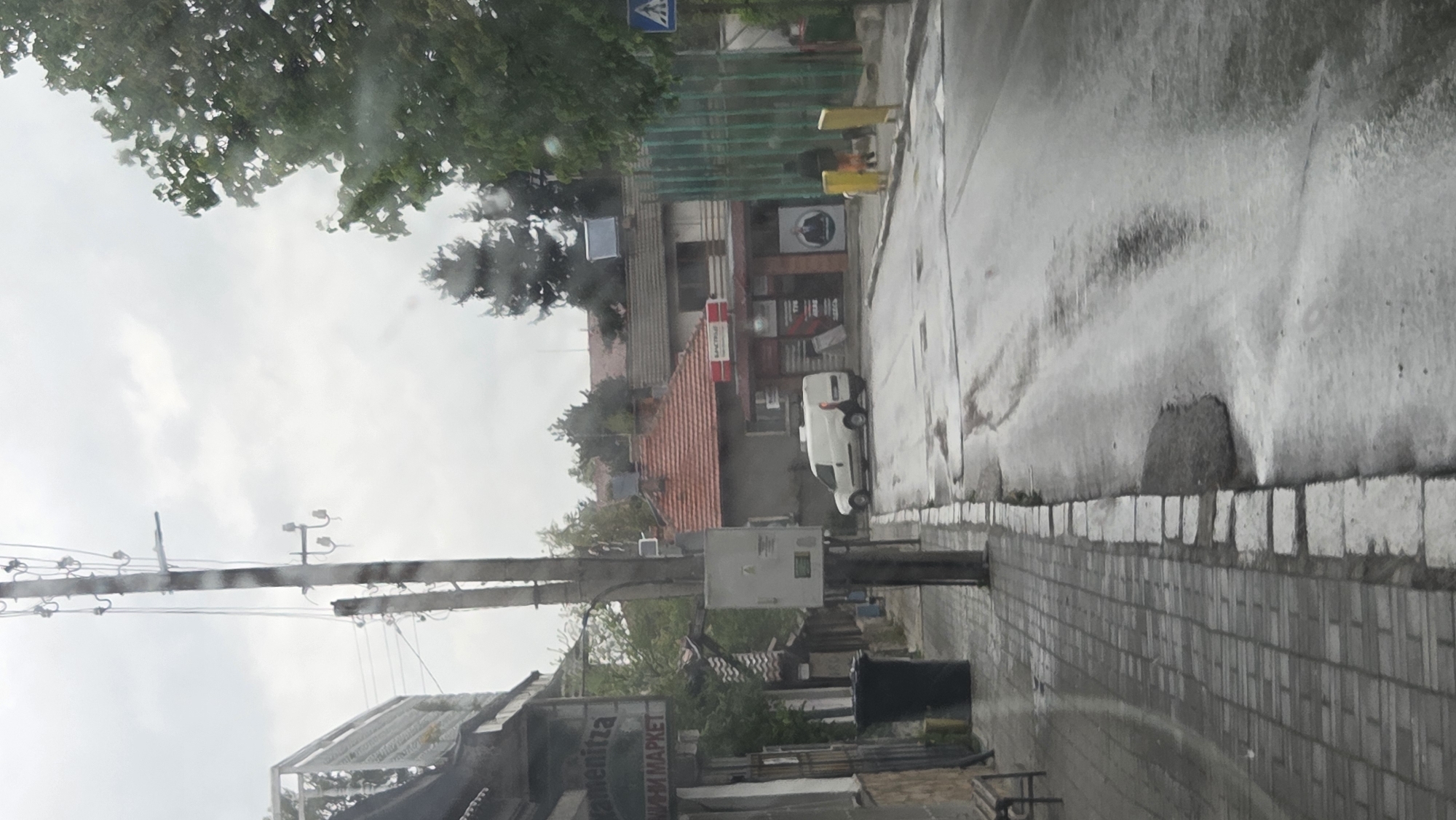 This media file presents a wet, overcast street scene, viewed from what appears to be a vehicle through a rain-streaked window. The image is rotated 90 degrees clockwise.
When viewed upright, the scene shows a street running horizontally, with buildings and trees on the left and a tiled sidewalk and power pole on the right. The wet surfaces of the road and sidewalk indicate recent rain or ongoing drizzle. The sky is uniformly grey and overcast, suggesting daytime.
**Subjects:**
* **Objects:** A prominent power pole with numerous overhead wires and an attached grey electrical box stands on the right side of the sidewalk. Further down the sidewalk, a black trash bin is visible. On the left side of the street, several buildings of varying architectural styles are visible, some with red-tiled roofs and others with corrugated metal siding. A white panel van is parked next to one of these buildings. Lush green trees are abundant, especially in the background and upper left. Yellow barriers or construction elements are present near the street.
* **Scene:** An urban or semi-urban street.
**Activity or event:**
No specific activity or event is taking place; it's a static street view.
**Notable details:**
* **Time of day:** Daytime, under a cloudy sky.
* **Weather:** The wet street, sidewalk, and visible moisture on the viewing surface (likely a car windshield) indicate recent or current rainfall.
* **Location cues:** A blue pedestrian crossing sign is visible in the upper left (when rotated). Visible text on signs, particularly "МАРКЕТ" (Market) and partially "СПЕЦИАЛ" (Special) in Cyrillic script, strongly suggests a location in an Eastern European or Balkan country.
**Visible text:**
* A blue square street sign with a white pedestrian crossing symbol.
* On a red sign: partially visible "СПЕЦИАЛ" (SPECIAL).
* On an advertising board: "МАРКЕТ" (MARKET) and other illegible Cyrillic letters, possibly part of "ОРМЕОНИТЗА" or similar.