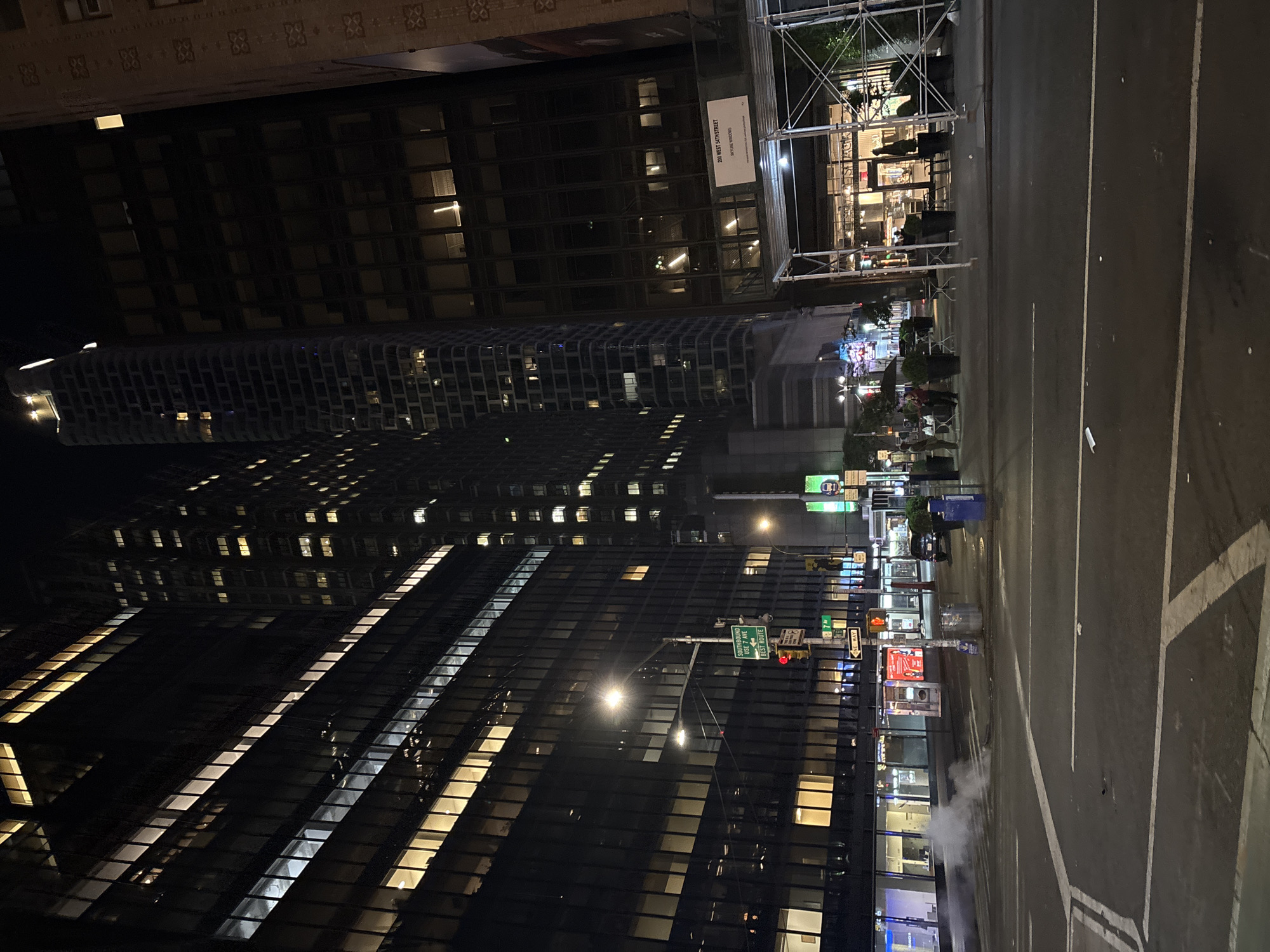The image presents a vibrant night-time street scene in a dense urban environment, confirmed by the location context as New York, United States of America. The perspective is from street level, looking down a thoroughfare flanked by tall buildings.
On the left, several high-rise buildings dominate the frame. Their dark glass facades reflect the city lights, with numerous windows illuminated in warm yellow and cool white, indicating interior activity. One building features a distinctive, grid-like architectural pattern, while an older structure with intricate stone facade details is partially visible at the upper left. A large banner on a building facade prominently displays the text "200 WEST 47TH STREET" and "SKYCORE WINDOWS", providing a precise location.
The street itself is paved asphalt with distinct white lane markings. A puff of steam rises from a grate on the right side of the road, a common characteristic of New York City streets. On the right, beyond the road, a sidewalk area is visible, featuring outdoor dining setups with tables, chairs, and planters. Extensive white scaffolding covers part of a building, beneath which the glowing facade of a "SHAKE SHACK" restaurant can be partially seen.
Numerous streetlights cast bright pools of light onto the road and buildings. Traffic lights, including a red one, along with various commercial signs, contribute to the nocturnal glow. Green street signs are visible, one clearly stating "47TH STREET ONE WAY" on its top line and "SOUTHBOUND" on the second line. Another sign below it appears to read "BEST ROUTE", and a yellow arrow sign indicates "ONE WAY". A few blurred figures are discernible on the sidewalks, including one person holding an umbrella, despite the absence of visible rain. The overall impression is one of a bustling, active city night.