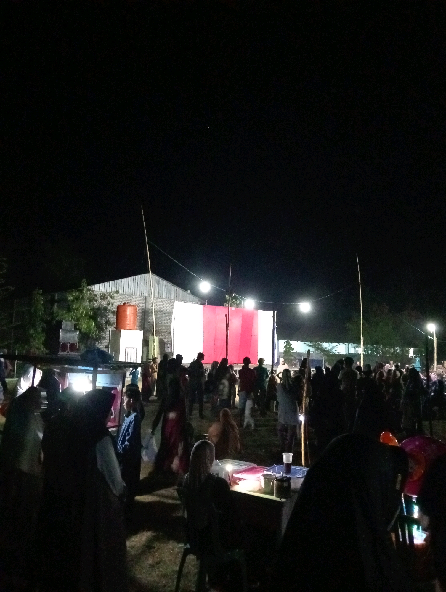 The image depicts an outdoor gathering at night in Pagerungan Kecil, Indonesia. A large crowd of people, silhouetted against lights, are present. In the background, a building with a corrugated metal roof is visible, along with a large orange water tank. A white and red banner is displayed across the front of the building, illuminated by lights. Several portable stalls or tables are set up, with some individuals interacting near them. The ground appears to be uneven and covered in grass or dirt. The overall atmosphere suggests a local event or festival.