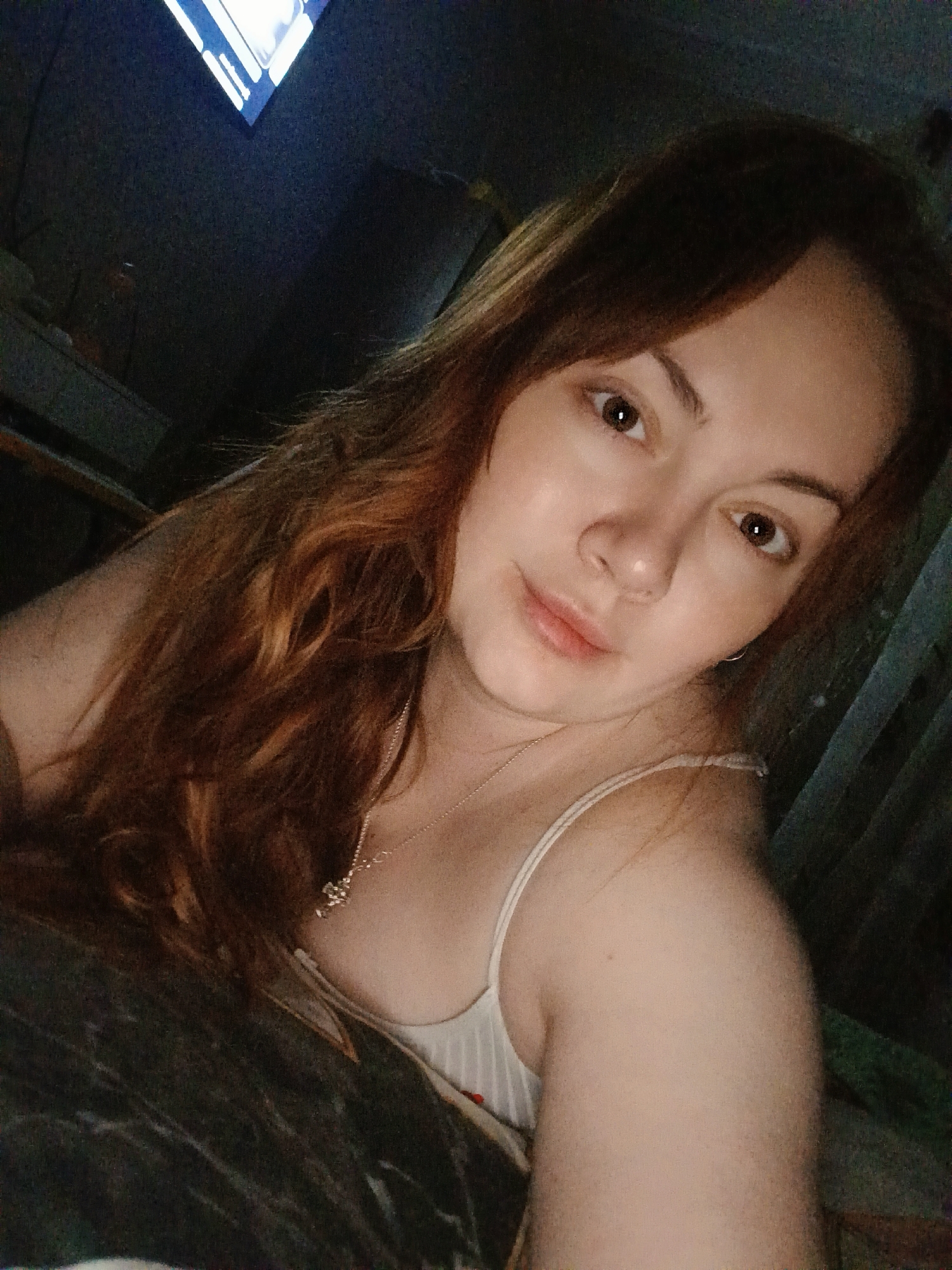 A woman with reddish-brown hair is taking a selfie. She is wearing a white tank top and a silver necklace. The background is dark and indistinct, but there appears to be a television screen visible in the upper left corner. The location is unknown, and there is no discernible evidence to indicate that the image was taken in the city of Chaikovsky, Russia.