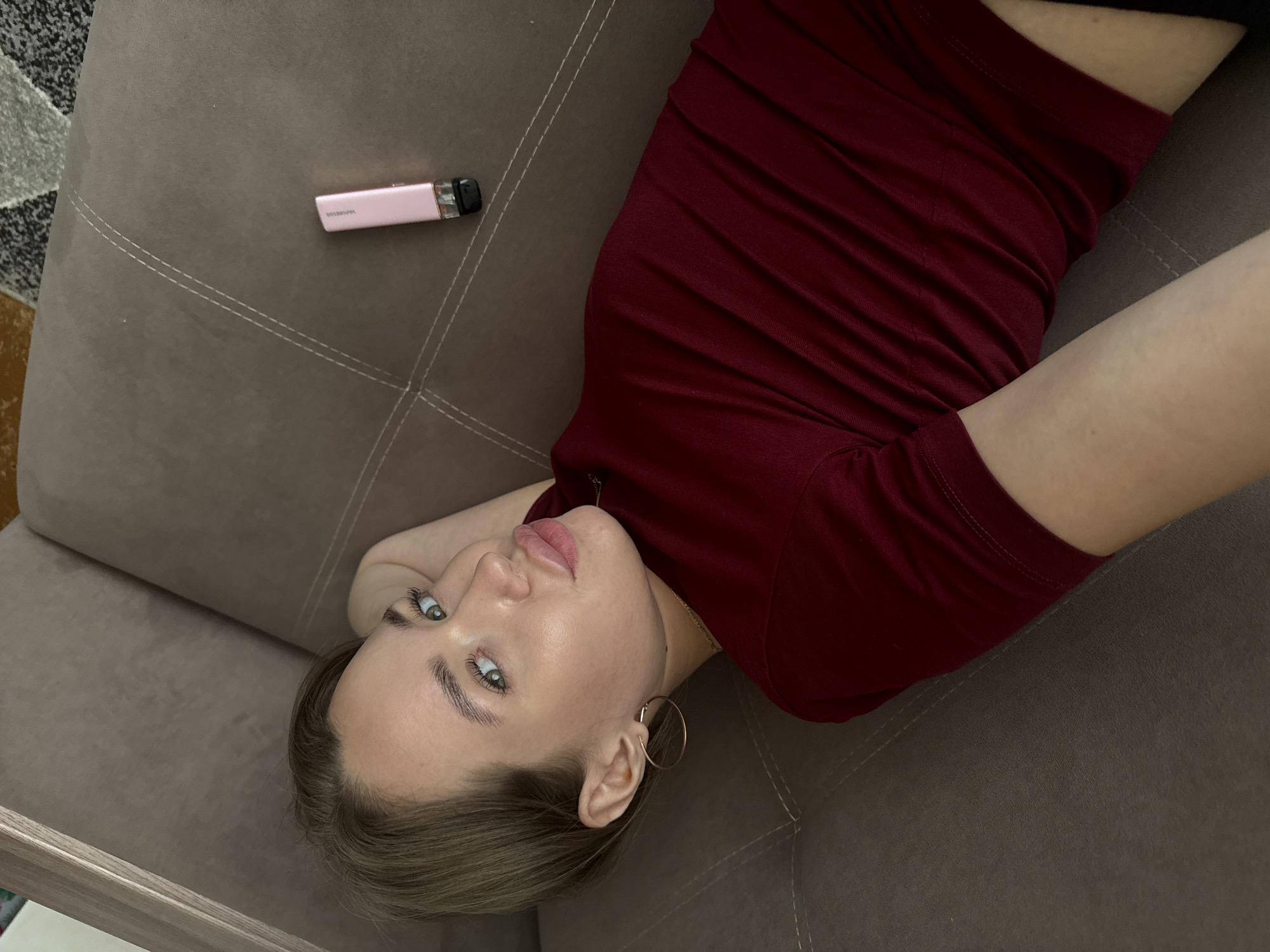 A young woman with short, brown hair and blue eyes lies on a brown sofa, looking at the camera. She is wearing a maroon t-shirt and large gold hoop earrings. Her left arm is propped behind her head. To her right, on the sofa, is a pink vape device. The sofa has visible stitching lines. The background consists of a patterned carpet. The lighting appears to be natural, coming from above and to the left. No specific details about the city of Chaikovsky, Russia are visible in the image.