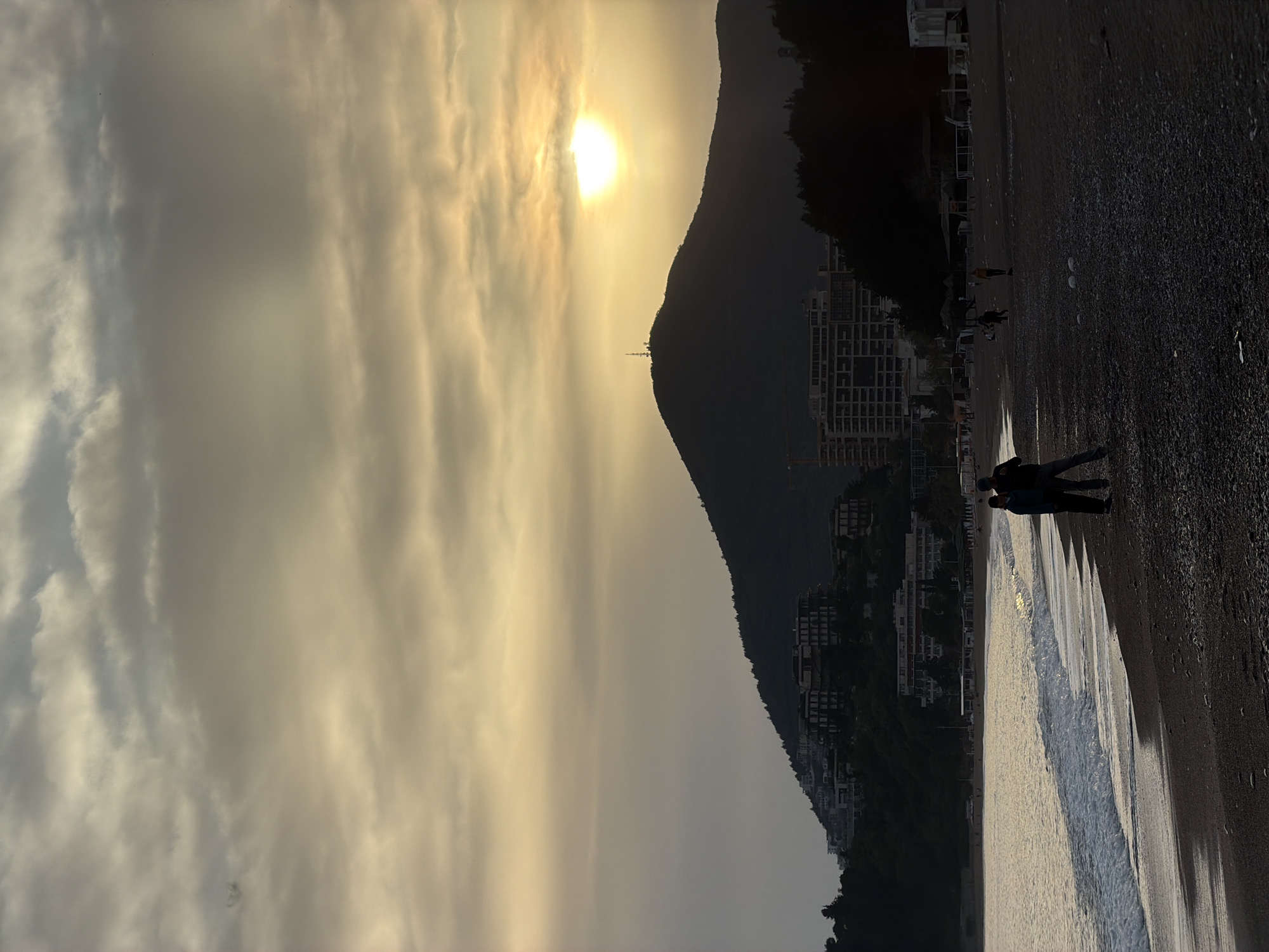 The image shows a beach in Rafailovići, Montenegro, at sunset. A large mountain dominates the background, obscuring much of the city. Buildings are visible on the slopes of the mountain. The sun is positioned behind the mountain, with a radio tower visible on the peak. Clouds cover the sky. Two people walk along the shoreline, their silhouettes facing the camera. The beach surface is a dark color. The incoming tide reflects the sunlight. Other people walk along the beach in the distance.