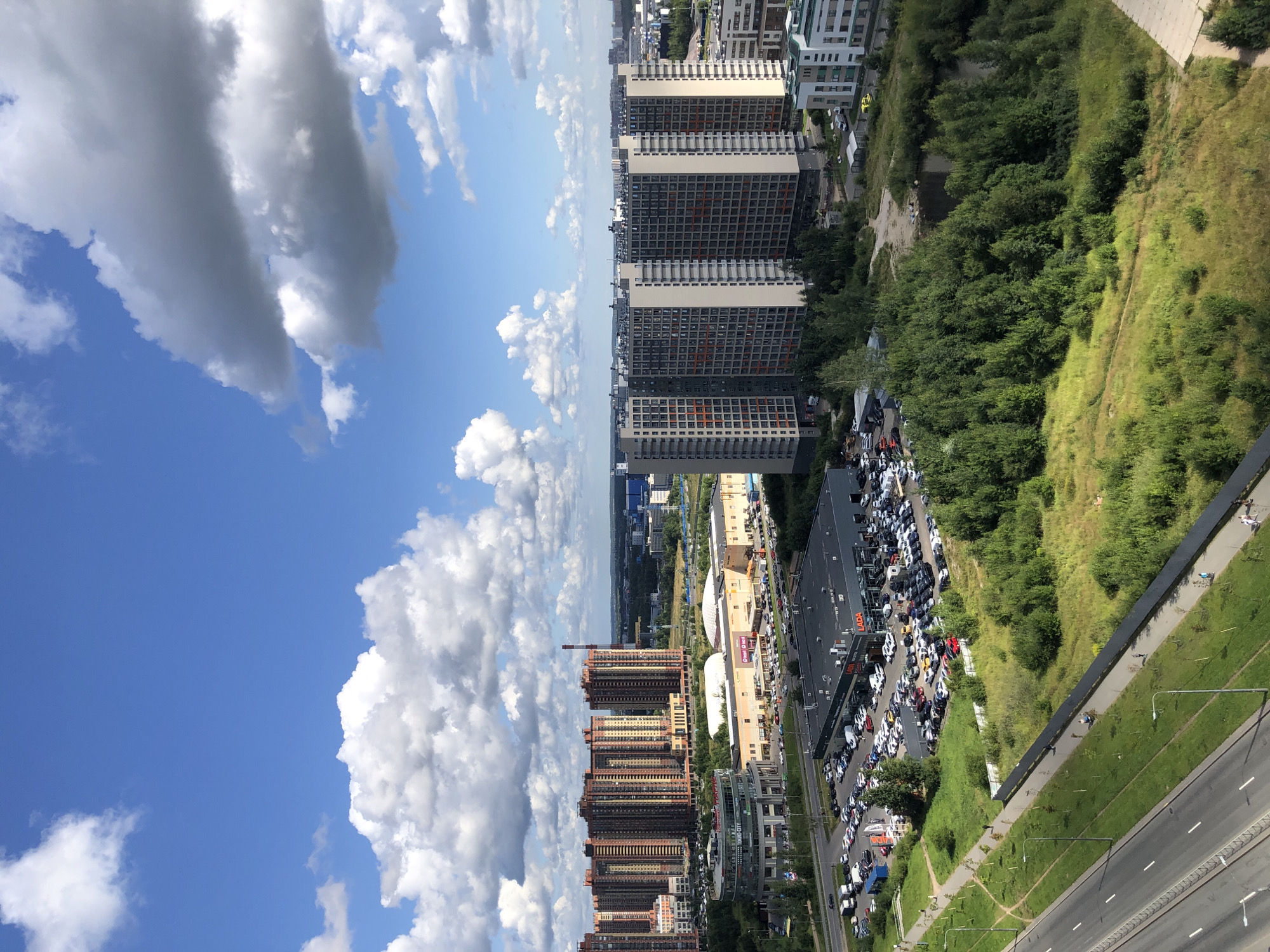 An aerial perspective captures a sprawling urban landscape under a bright blue sky dotted with white, cumulus clouds. In the foreground, a multi-lane road runs diagonally across the frame, bordered by a grassy median and a sidewalk. Further back, a vast, green field stretches out, interspersed with trees and a pathway. Adjacent to the field is a large parking lot filled with numerous cars, situated in front of a dark-colored, rectangular building with a "LADA" sign prominently displayed.
Beyond the parking lot, a cluster of modern, multi-story residential buildings with distinct architectural styles and colors dominate the mid-ground. To the left, a row of tall, reddish-orange apartment buildings stands out, while to the right, a group of taller, gray and beige buildings with visible orange accents forms a more uniform block. In the distance, a hazy horizon reveals more city structures and industrial elements, including a tall, red-and-white striped chimney. A few smaller buildings and green spaces are also visible throughout the scene. The overall impression is one of a developed, modern urban area on a clear, sunny day.