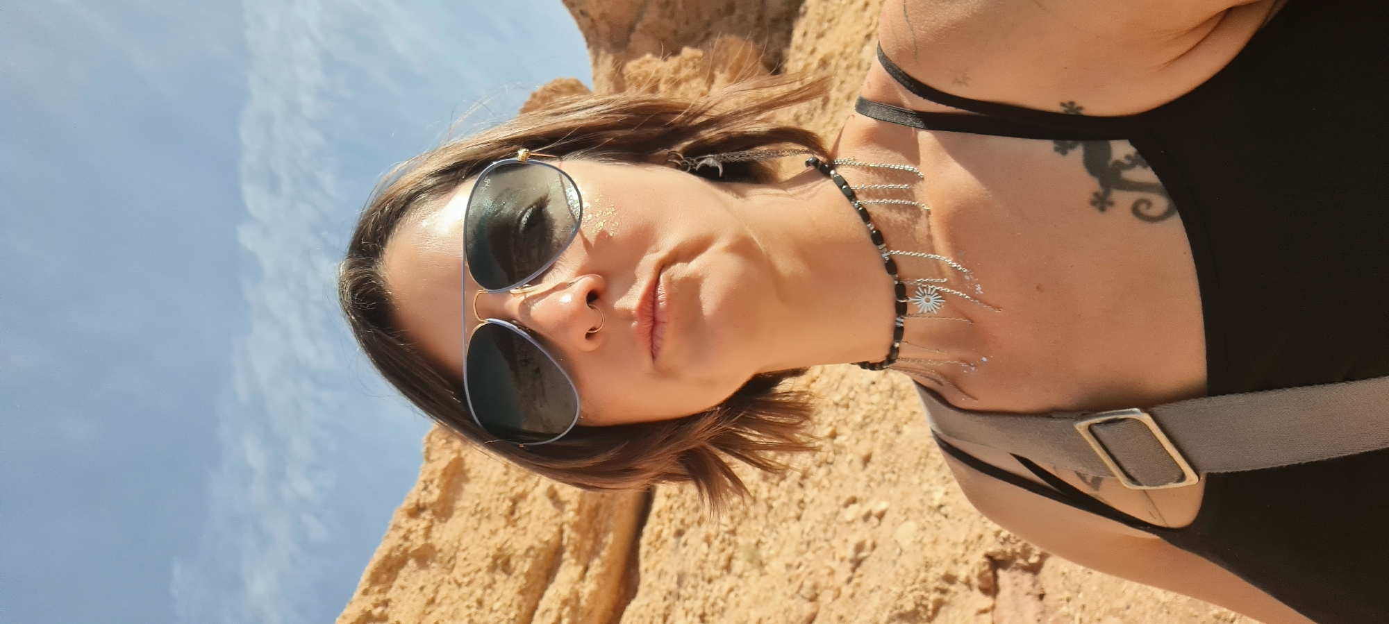 A medium shot angled up at a woman with short, brown hair is presented. She is wearing sunglasses with blue frames, a black tank top, and a grey strap across her shoulder. She has a nose ring and a delicate black and silver necklace with a small flower charm. A lizard tattoo is visible on her upper left shoulder. The background consists of a sandy or rocky landscape and a partly cloudy blue sky. The location appears to be in a desert area within the Hevel Eilot Regional Council, Israel.
