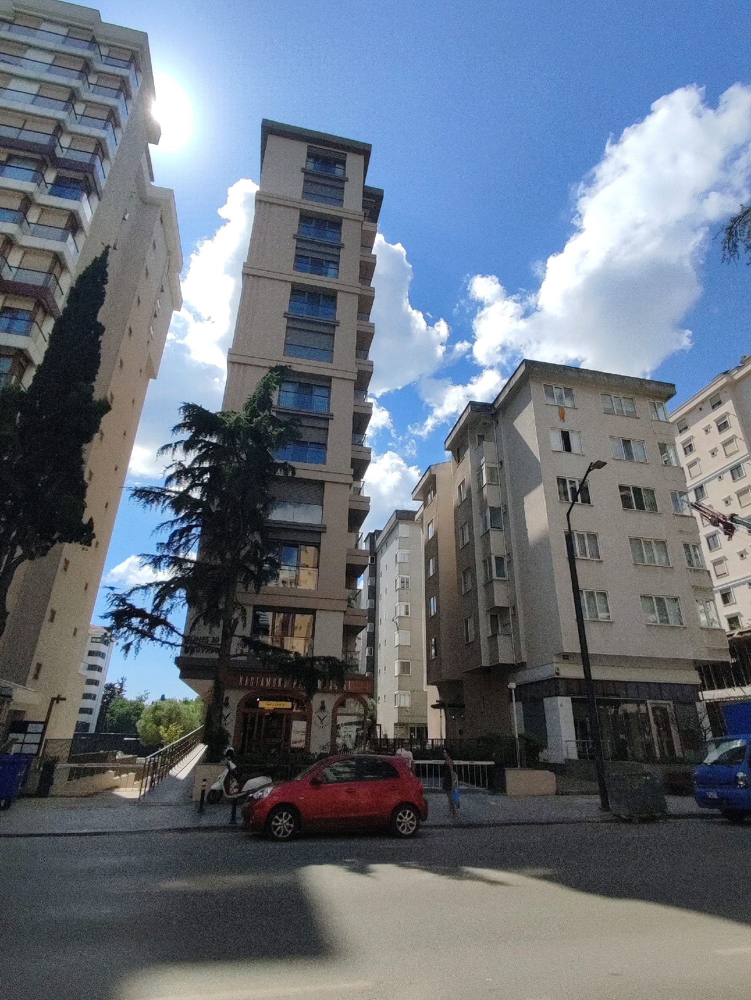 The image depicts a street scene in Kadıköy, Turkey, characterized by a dense urban environment with several multistory buildings. In the foreground, a red hatchback car is parked on the right side of the asphalt road. To the left of the car, a person in a blue shirt and light-colored shorts is standing near a white metal barrier. Behind the barrier, there is a walkway leading up to a building with a storefront displaying "Kastamonu Pastanesı."
Dominating the background are three tall, modern buildings of varying designs. The tallest building, a slender tower, is located centrally and is beige in color with numerous windows. To its left is another tall, lighter-colored building with visible balconies. To the right of the central tower is a shorter, grey building with a more traditional apartment block appearance, featuring many windows. The sky is bright blue with scattered white clouds, and sunlight is strong, casting shadows on the road. Trees and greenery are visible between the buildings, adding touches of nature to the urban landscape. The overall impression is of a typical day in a busy Turkish city.