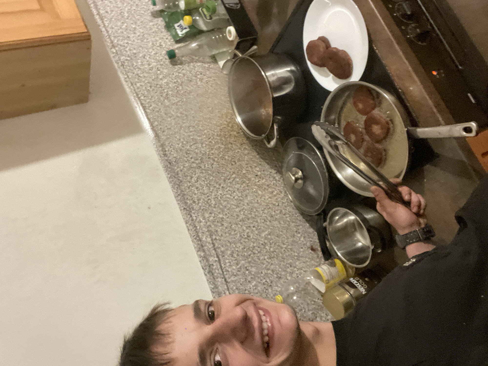 This scene takes place in a domestic kitchen in the city of Unknown, Unknown.
A male individual, appearing to be a young adult, is captured in a selfie. He is smiling broadly, directly facing the camera with his brown eyes. His short, dark brown hair has bangs across his forehead. He wears a black long-sleeved shirt and a dark-colored watch on his left wrist. His right hand holds metal cooking tongs, positioned above a frying pan on an electric stove.
The stove has multiple burners; the front panel is dark-colored with control knobs and a small red indicator light illuminated, indicating it is active. The frying pan contains a liquid (likely cooking oil) and four round, dark-brown, disc-shaped food items (resembling patties or fritters) which are actively cooking, with visible bubbling. To the right of the frying pan, a white round plate holds two identical, cooked, dark-brown disc-shaped food items. On another burner to the left, a large stainless steel pot sits, with its lid slightly ajar, and steam is visible rising from within. A glass pot lid with a metal handle rests on the countertop to the left of the frying pan.
The countertop and backsplash are made of a light-colored, speckled material. Various items are on the counter: a gold-colored jar labeled "NESCAFÉ Gold," a yellow-capped clear plastic bottle, a small empty metal bowl, and several plastic bottles (some clear, some with green caps, one with a "Just" label) housed in a black tray or holder in the upper right portion of the image. The wall above the backsplash is plain light beige/off-white, and a light-wood kitchen cabinet with a panel door is visible in the upper right corner.
