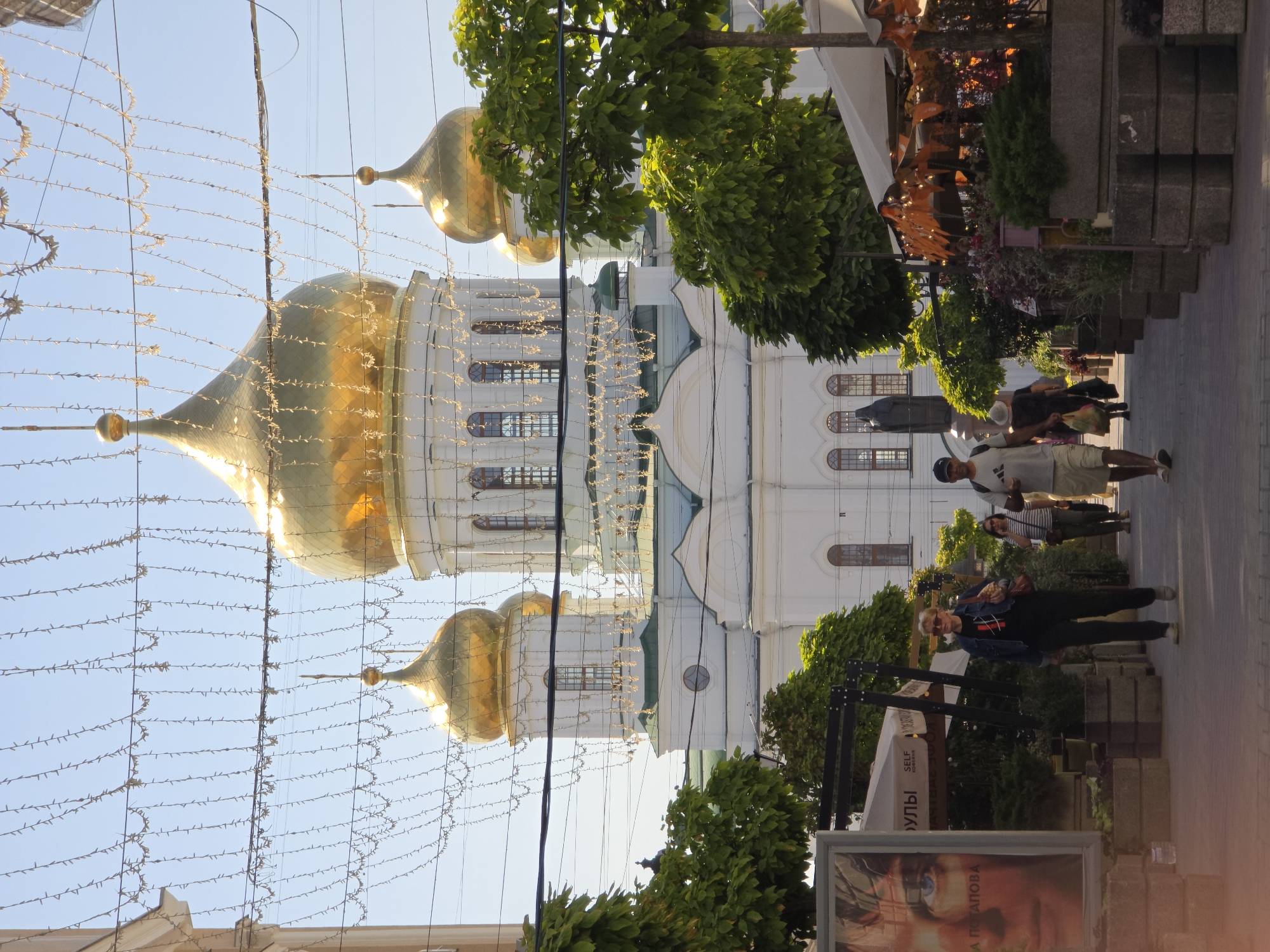 A street scene in Rostov-on-Don, Russia, features a large white building with golden domes, identified as the Nativity Cathedral, dominating the background. The building has multiple windows and architectural details. In the foreground, a pedestrian walkway is lined with trees, planters, and outdoor seating areas for cafes, some with umbrellas and decorative lights. Several people are walking along the path. A large poster of a person's face is visible on the left side of the image. Overhead, strings of lights are strung across the street. The sky is clear and blue, indicating a sunny day.