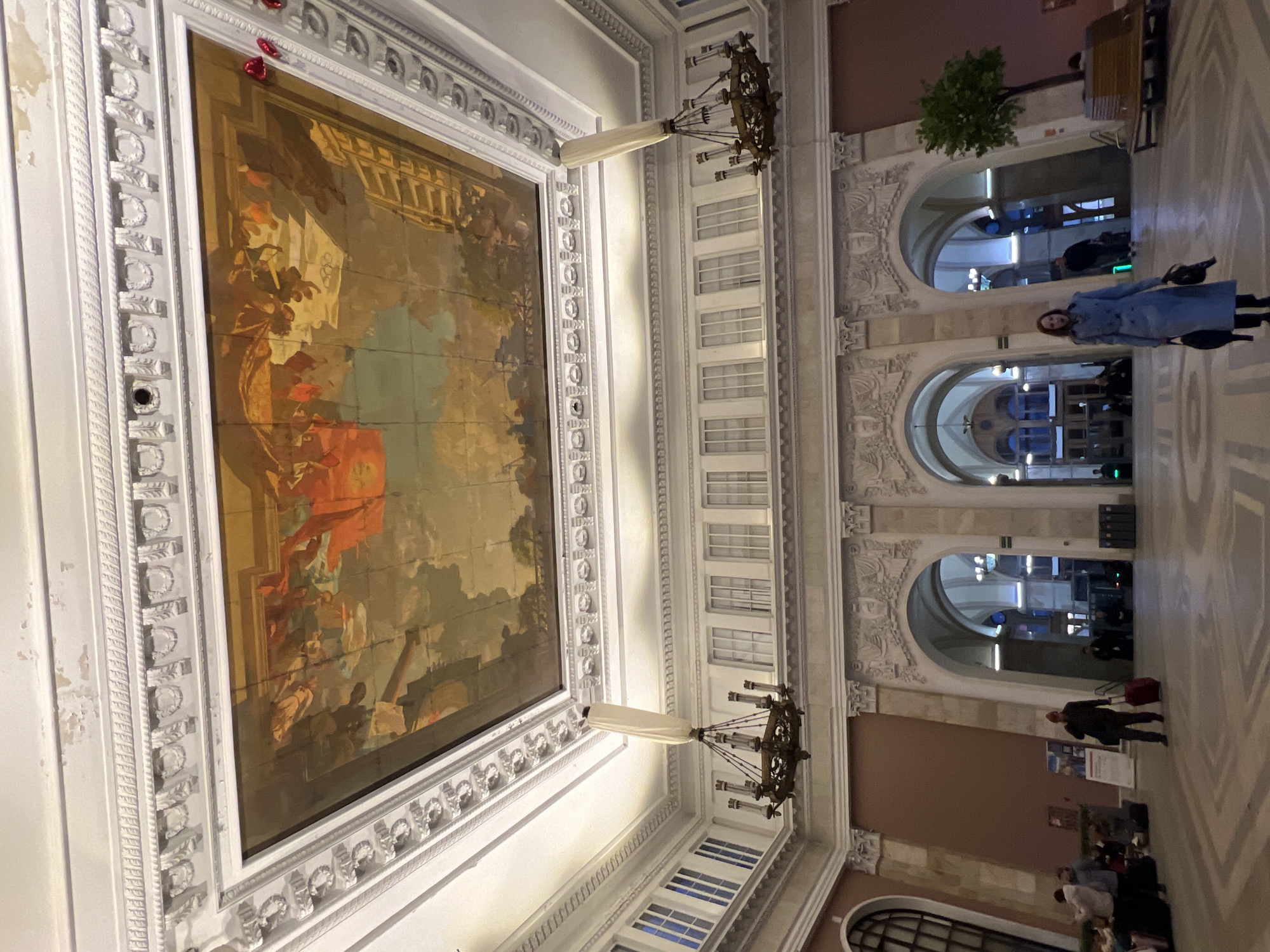 The image depicts the interior of Vitebsky railway station in Saint Petersburg, Russia. The ceiling is ornate, featuring a large rectangular painting framed by white molding. Two chandeliers are suspended from the ceiling. The architecture includes archways and detailed plasterwork. People are walking through the station. A woman in a blue coat is visible in the foreground. The floor has a geometric pattern.