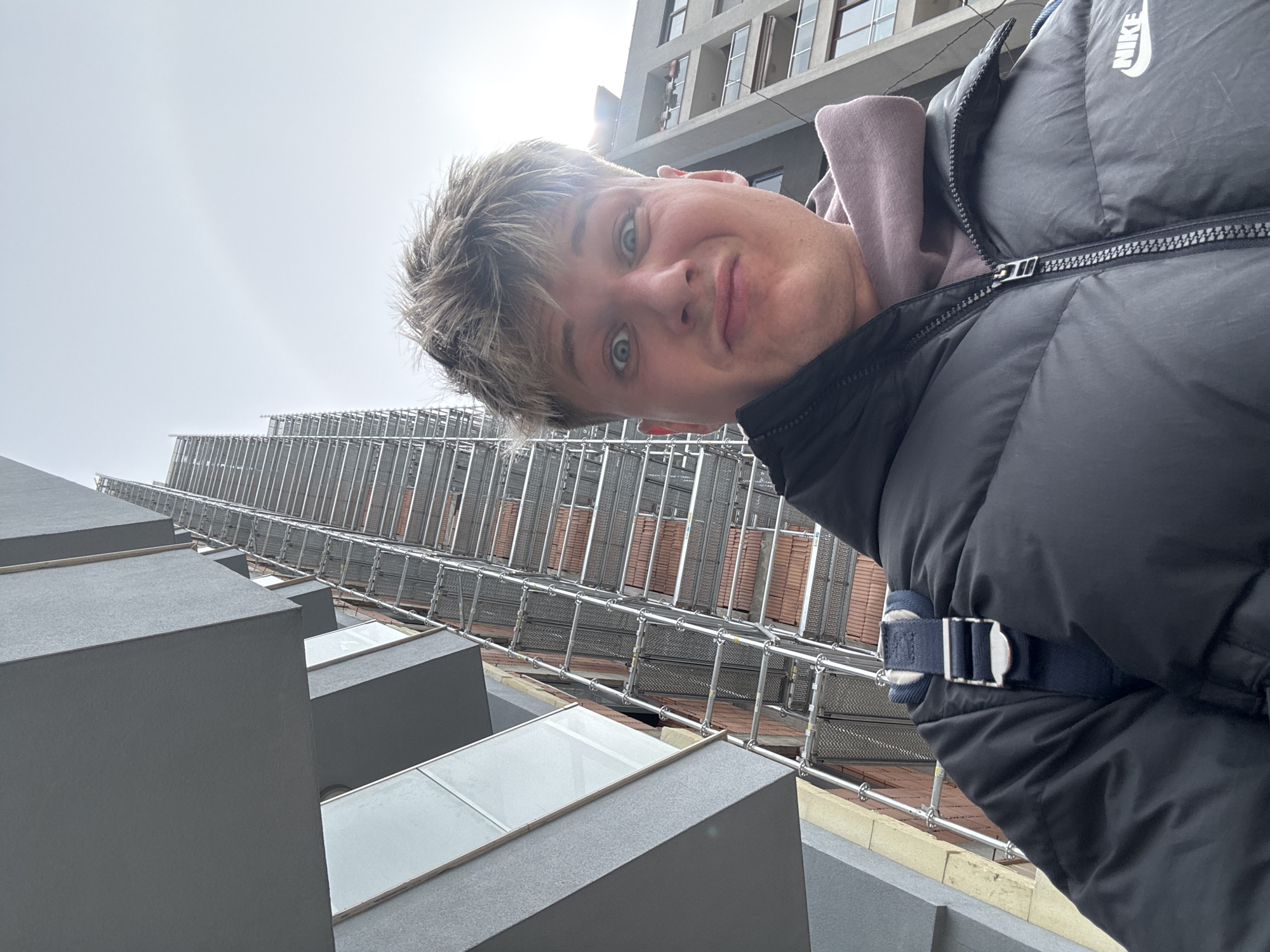 A selfie taken from a low angle shows a young, light-skinned man with short, blond-highlighted hair and blue eyes, wearing a gray hooded sweatshirt and a black Nike puffer jacket. He is looking down at the camera with a playful expression. Behind him, a tall building is visible, covered in scaffolding. The building appears to have a modern architectural design with gray concrete or similar material forming protruding rectangular shapes, some of which have glass windows. The sky is overcast and bright. While I don’t have specific location data in Plovdiv, Bulgaria, the architecture and scaffolding could indicate urban construction or renovation work.