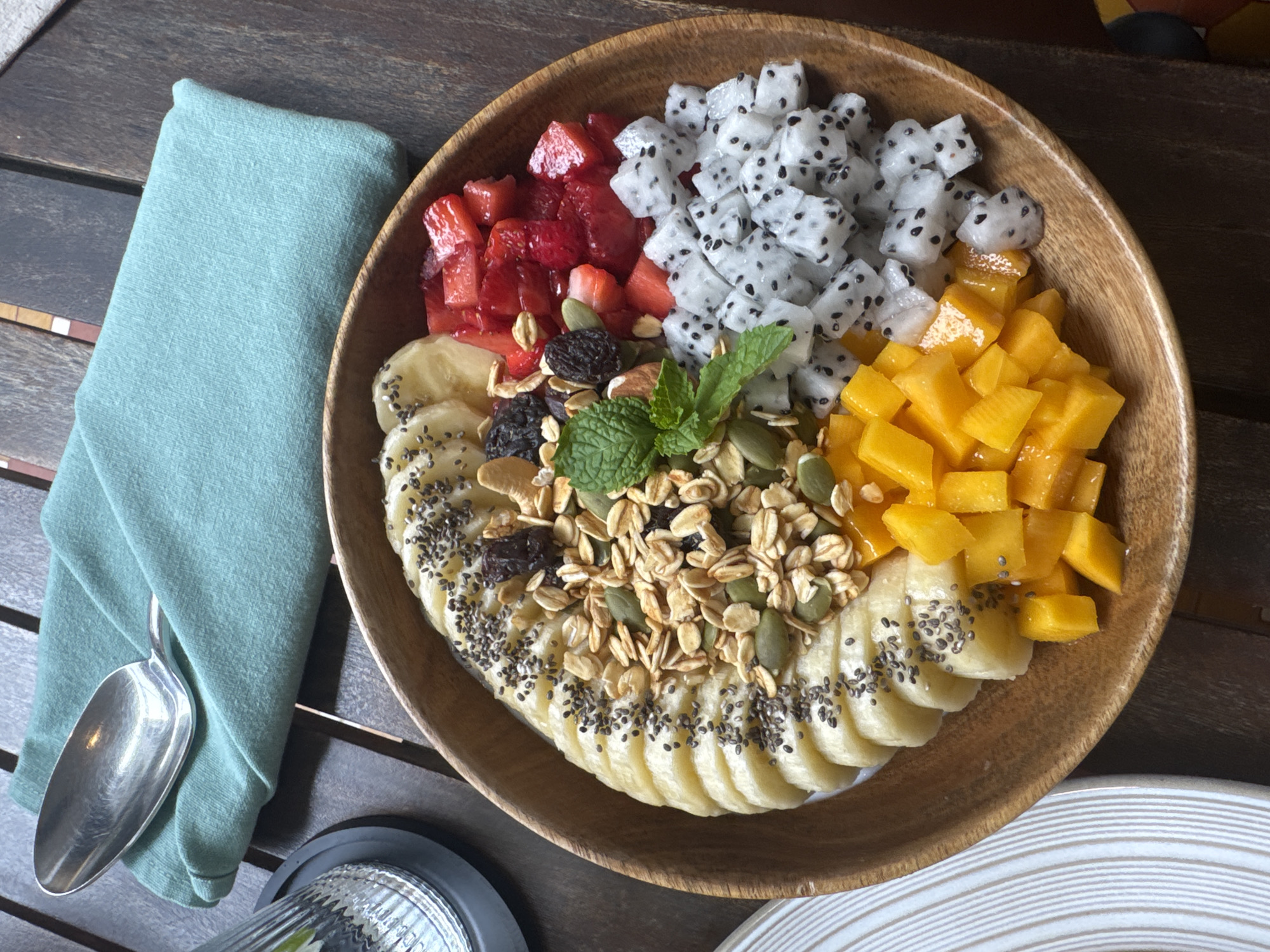 A top-down, eye-level, close-up shot shows a wooden bowl of fruit sitting on a wooden table next to a turquoise napkin with a spoon. The bowl is filled with sliced bananas topped with chia seeds and granola, diced strawberries, diced dragon fruit, and diced mango. A sprig of mint is placed in the center. The scene is presumed to be located in Đà Nẵng, Vietnam based on the visual evidence. No people are visible.