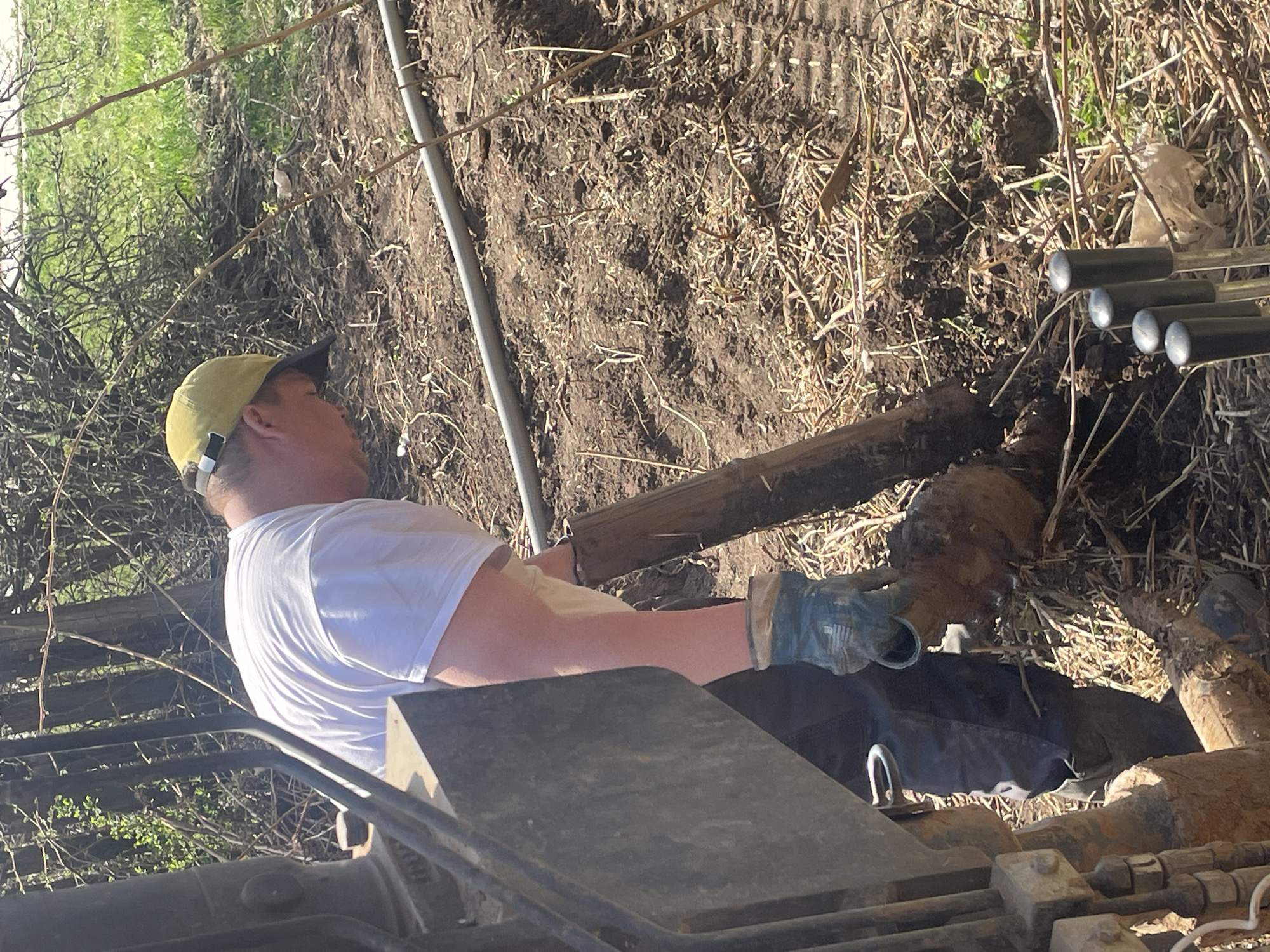 A construction worker in Поедуги, Russia, wearing a yellow cap, white t-shirt, and work gloves, is operating a piece of heavy machinery, possibly for excavation or earthmoving. The worker is holding a muddy log. The environment appears to be a construction site with exposed dirt and scattered debris, including pipes. Vegetation, possibly overgrown, is visible in the background.