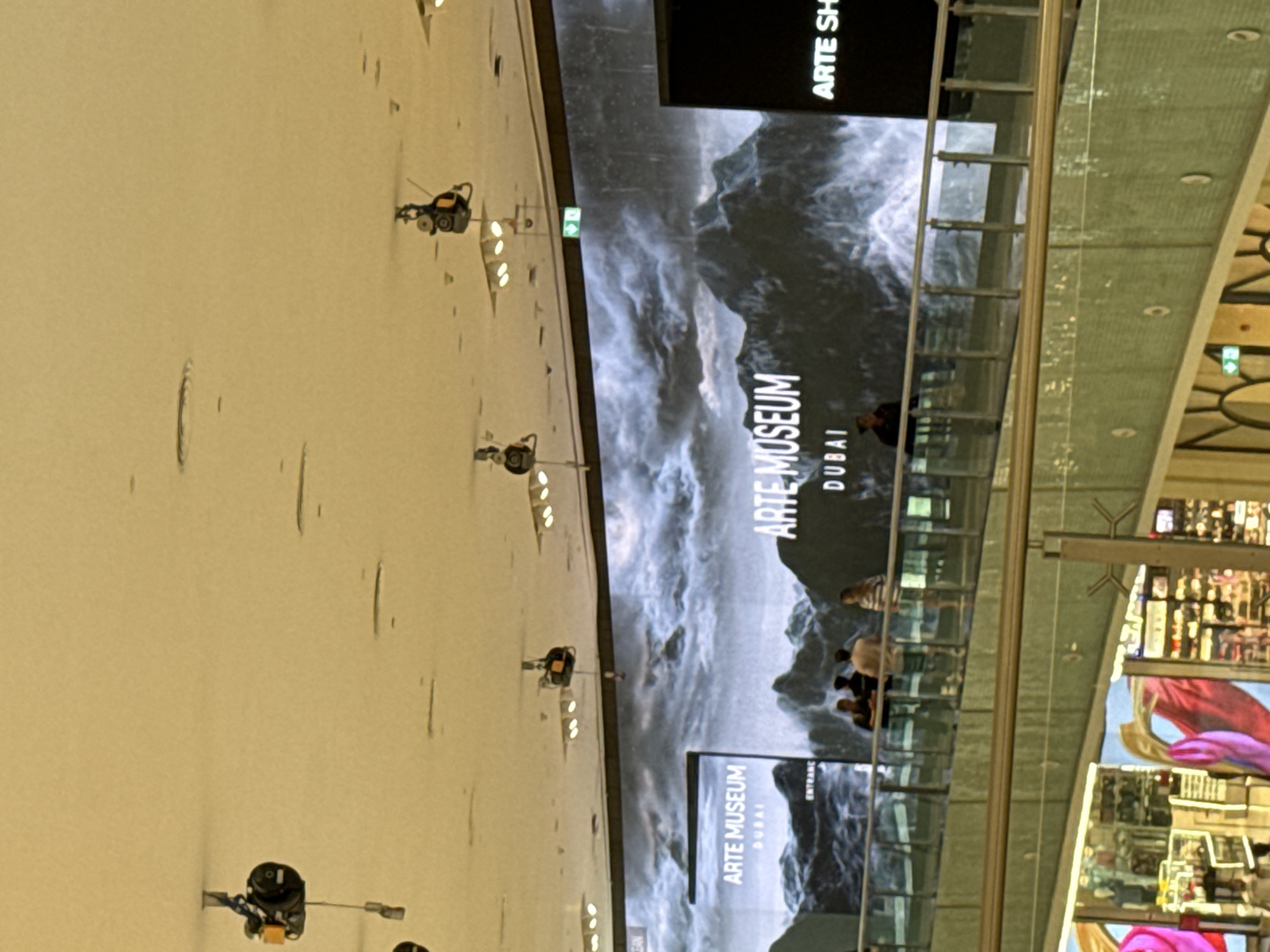 The image shows a large digital screen displaying "ARTE MUSEUM DUBAI" with a backdrop of mountainous scenery. Several spotlights are visible on the ceiling. Below the screen, a railing separates a walkway where a few people are standing and observing. The lower section of the image reveals shops and displays on a different level of the building, suggesting it is a large indoor venue, likely a mall or exhibition space in Dubai, United Arab Emirates.