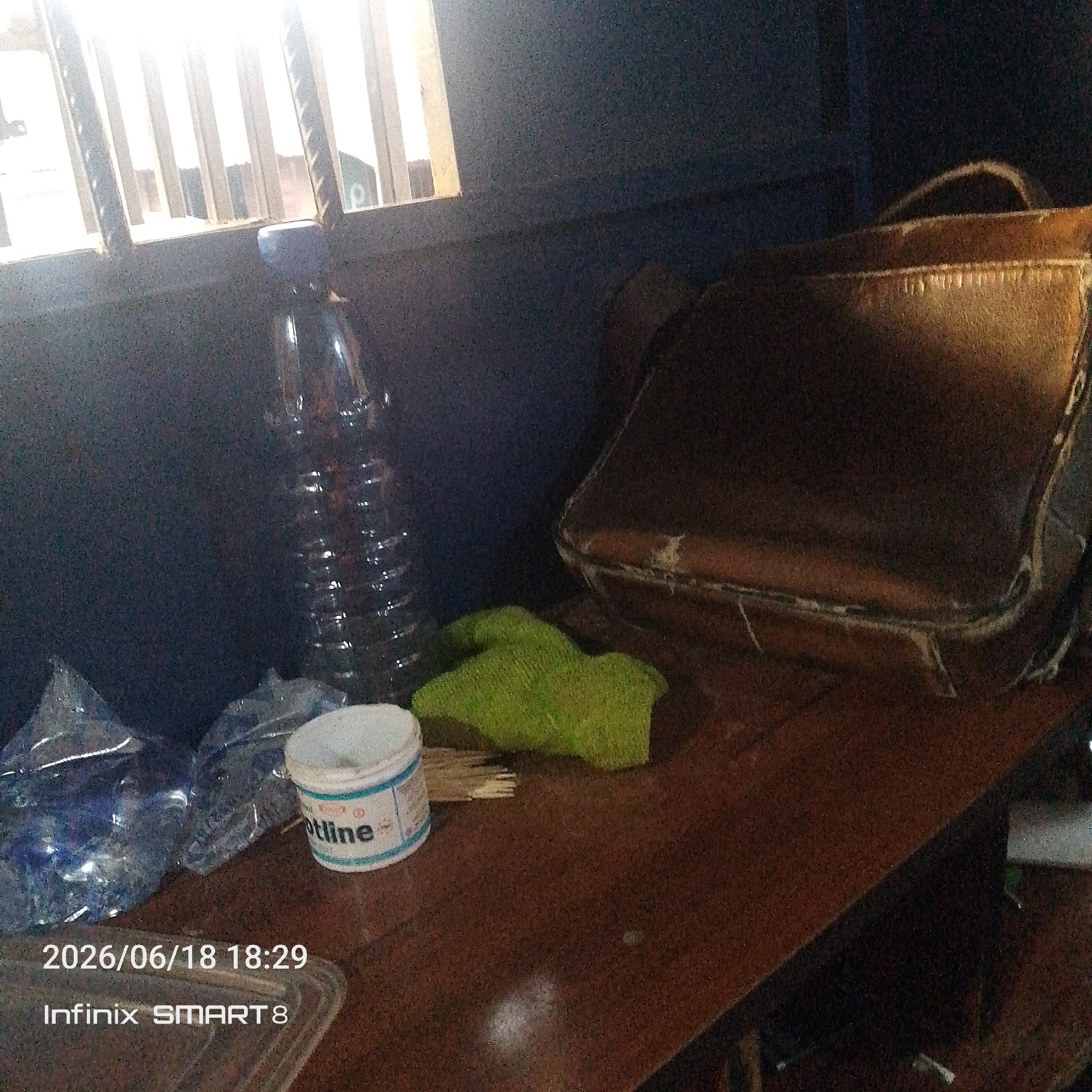 This media file depicts a dimly lit indoor scene. The primary subjects are a collection of objects on a wooden surface, likely a table or desk. In the foreground, to the left, are several plastic bottles, some appearing to be partially filled with a blue liquid. Next to them is a small white tub with a blue label. Text on the tub is partially visible and reads "tline". To the right of the bottles, a green, textured object is placed, possibly a cloth or a sponge. Further to the right and behind these items is a dark brown, leather bag with a curved handle. The bag appears worn and has visible stitching.
In the background, a window is visible with vertical slats, allowing bright light to stream through, creating strong highlights and shadows across the scene. The overall atmosphere is somewhat cluttered, with the objects arranged somewhat haphazardly.
Visible text at the bottom left of the image indicates the date and time as "2026/06/18 18:29" and the device used as "Infinix SMART 8". No people are visible in the frame. The lighting suggests it could be late afternoon or evening, with artificial light supplemented by the natural light from the window. The location is indeterminate, but the presence of the bag and the window might suggest an indoor workspace or a home setting.