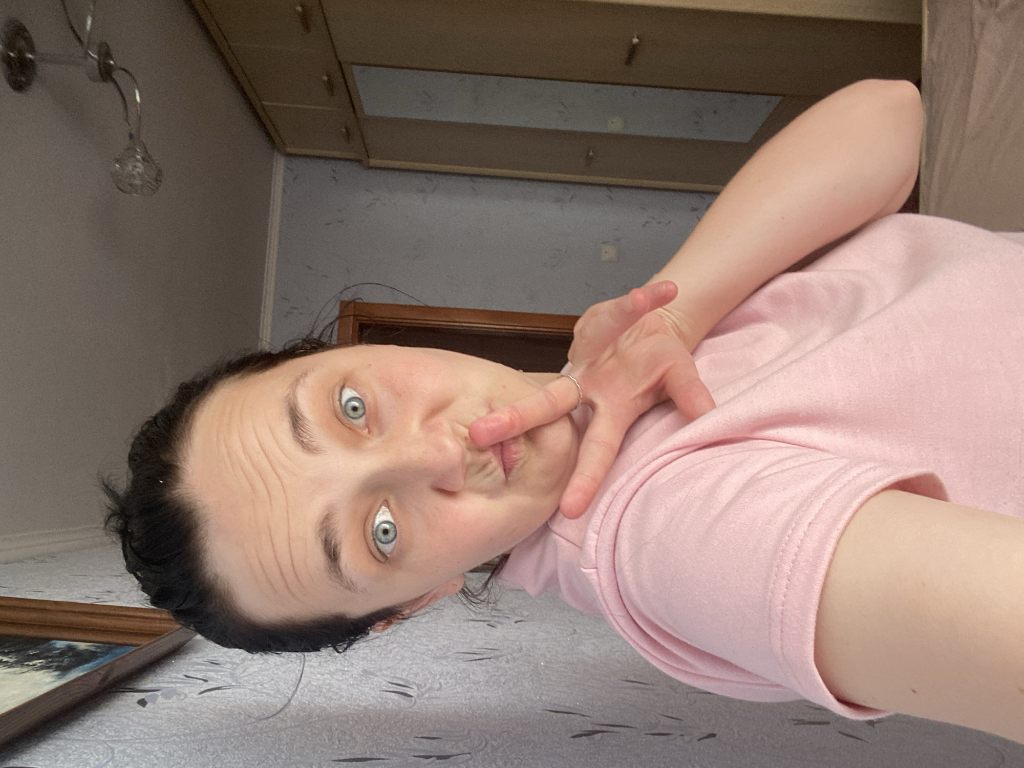 A selfie taken by a woman in her 20s, wearing a pink t-shirt and has dark hair pulled back, is looking at the camera with a playful expression, her finger on her lips as if to shush the viewer. Her eyes are wide and blue. Her right hand is held up, forming a peace sign. The background shows a wall with patterned wallpaper, a framed picture on the left, a closet with mirrored doors on the right, and a ceiling light fixture. The location is indoors in what appears to be a bedroom.