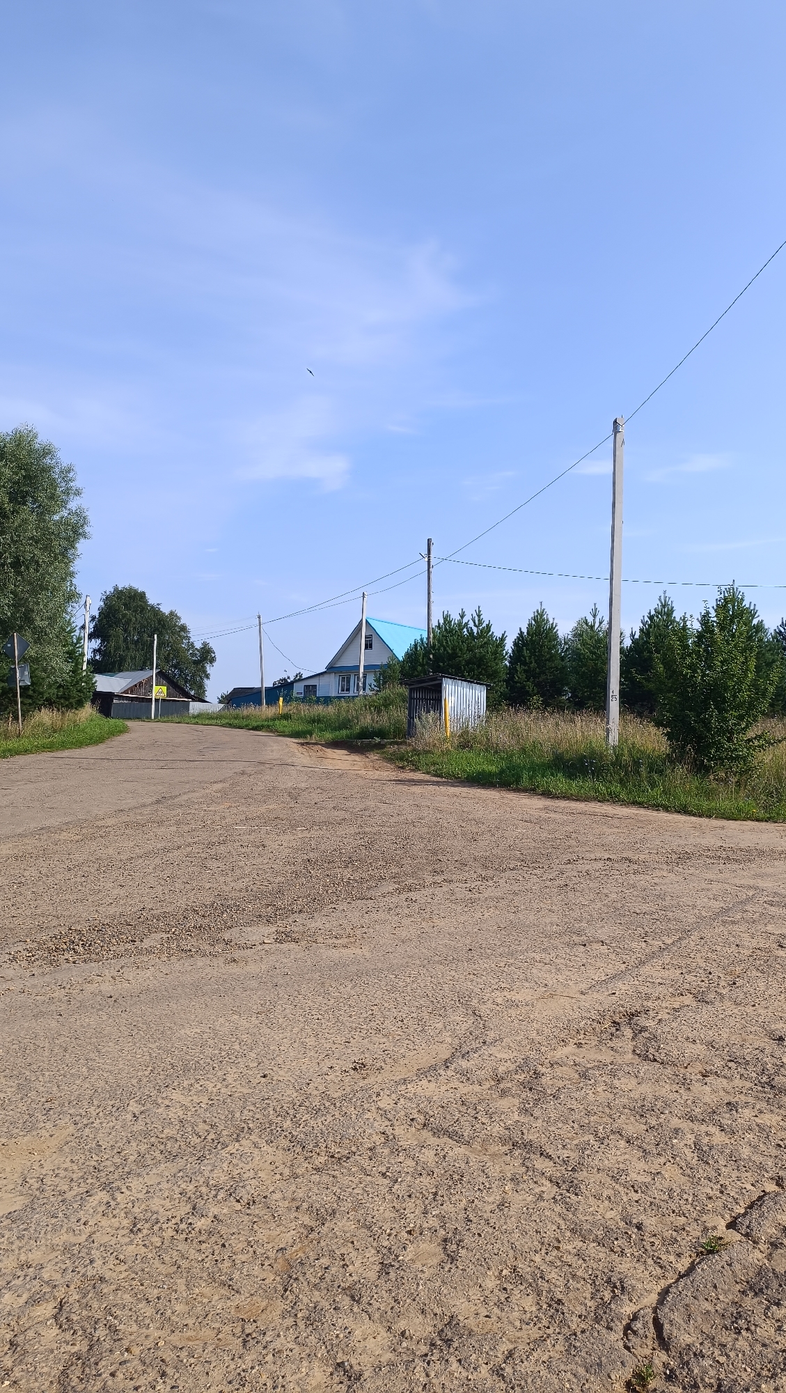 The image shows a dirt road leading towards a rural settlement under a clear blue sky. On the right side of the road, there is a metal shed with yellow markings. Further along the road, a light blue roof of a house is visible, surrounded by trees and vegetation. Utility poles with wires stretch across the scene. In the distance, a small bird can be seen in the sky. The road surface is rough and uneven, indicating it is unpaved. There are no people or animals visible in the image. The setting appears to be a rural landscape in Russia.