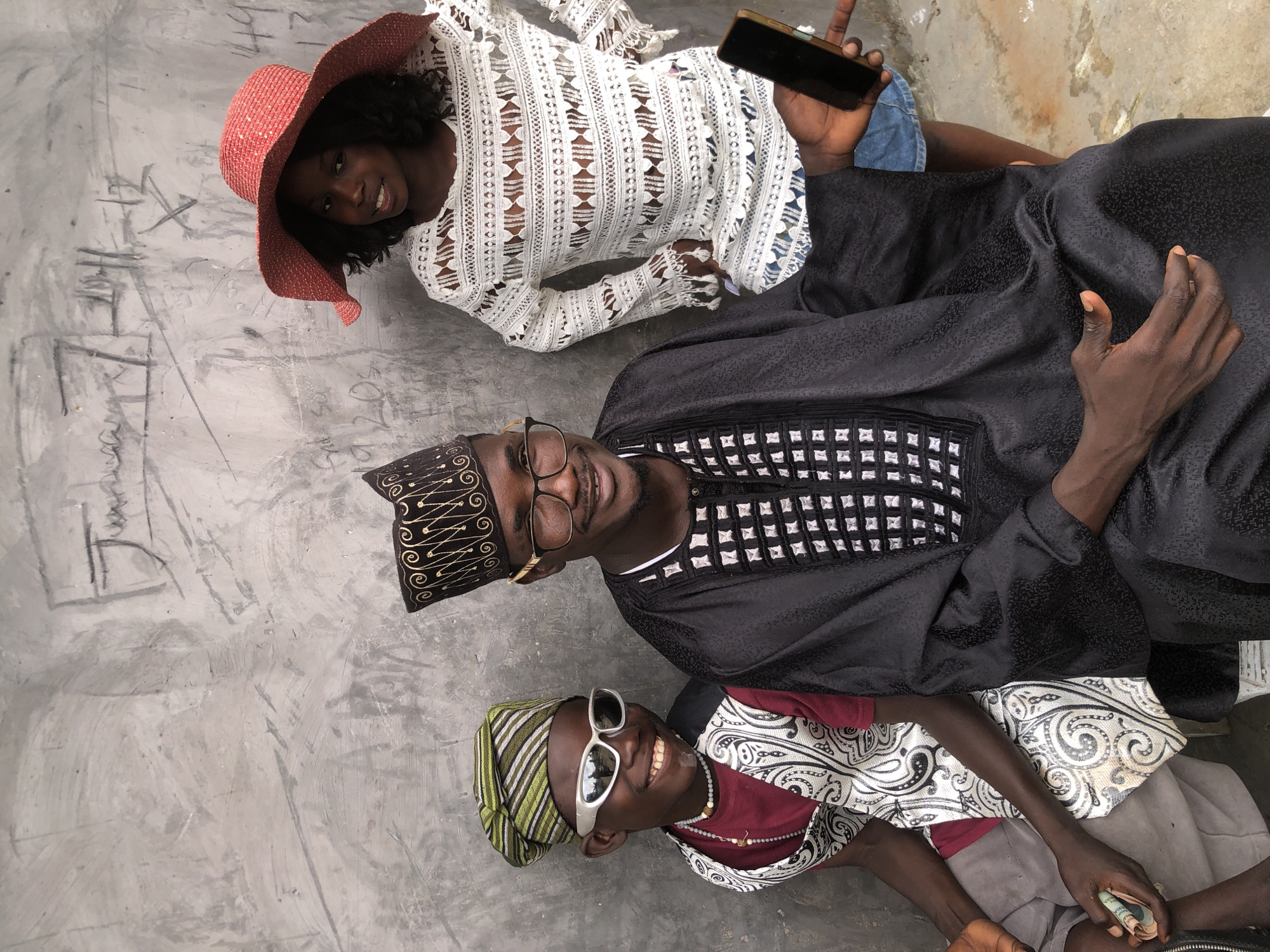 This image captures three individuals posing against a textured, grey concrete wall in Ado, Nigeria. The wall itself is marked with visible graffiti and scuff marks.
On the left, a young boy is seated and facing forward, sporting white-framed sunglasses and a green and white striped cap. He wears a dark red t-shirt and a white vest with a black and white patterned design. A beaded necklace is visible around his neck. He is holding a small roll of cash in his left hand.
In the center, a man is seated and looking towards the camera with a smile, wearing glasses and a black cap with gold embroidery. He is dressed in a black robe adorned with a white and black patterned design on the chest. His right hand is extended, holding a black smartphone.
On the right, a young girl stands, facing slightly away from the camera but looking towards the viewer with a smile. She wears a wide-brimmed red hat and a white, intricately patterned long-sleeved top over what appears to be denim shorts.
The lighting suggests it is daytime, possibly overcast, with soft illumination. The overall scene appears casual and posed.