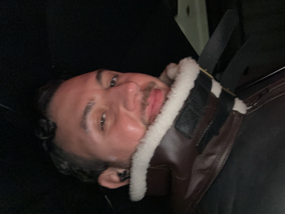 A man with dark hair and a light complexion is pictured close-up. He has a slight mustache and a small amount of stubble. He is wearing a brown leather jacket with a white, furry collar. The photo appears to be taken from a low angle, with dark surroundings suggesting an indoor setting. Safety seatbelts are visible above his right shoulder.