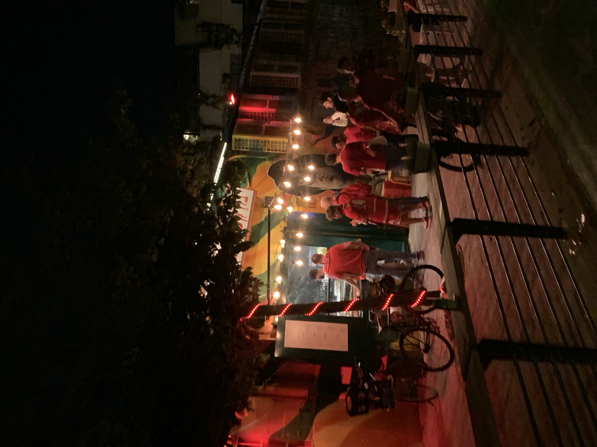 The scene depicts an outdoor establishment in Tel Aviv, Israel, at night. Several people are gathered under string lights, some wearing red shirts, suggesting a possible event or gathering. A menu board is visible near a bicycle with red lights. The environment includes a building with multiple windows and a fence along a walkway.
