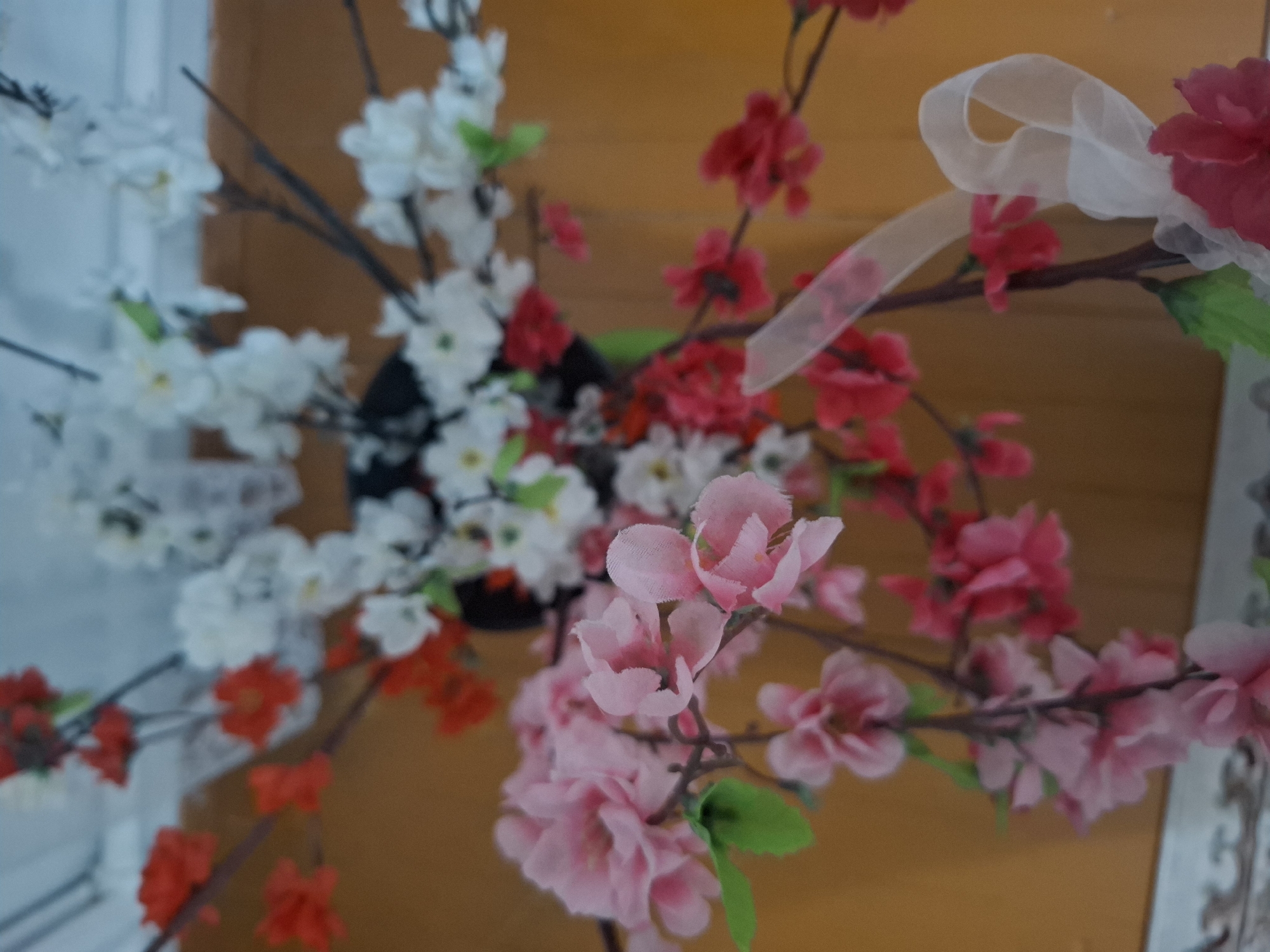 The image displays a close-up, overhead view of artificial flowers on branches. The flowers are a mix of white, pink, and red, with some green leaves visible. A sheer white ribbon is tied to one of the branches. The background is a warm, orange-brown color, possibly a wall or furniture. The scene appears to be indoors, with a hint of a light blue ceiling in the upper portion of the image. There are no people or discernible actions taking place. The location of this image within Almaty, Kazakhstan cannot be determined from the visual information provided.