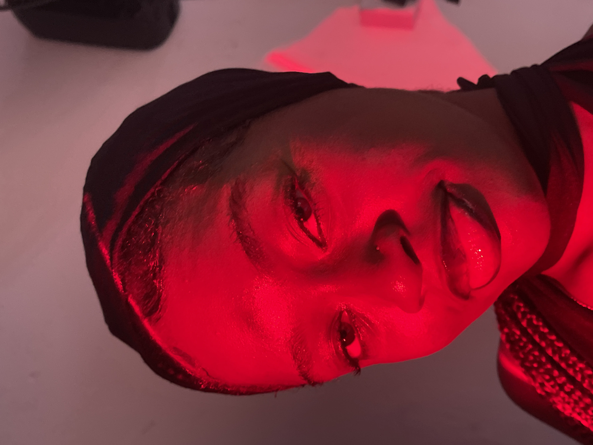 The image presents a close-up, artistic portrait of a person, likely female, illuminated predominantly by an intense red light. The subject's face is central, depicted from a low, slightly upward angle, with the head tilted to the right. The strong red light casts vibrant hues across the skin, highlighting the contours of the eyes, nose, and lips, and creating dramatic shadows. The person's eyes are open, looking upwards or slightly away, conveying a contemplative or pensive expression. The lips are slightly parted.
A dark head covering, possibly a scarf or durag, frames the face. On the right side of the image, near the person's neck and shoulder, a textured material with a woven pattern, also bathed in the red light, is visible. This could be part of their attire or hair.
The background is softly blurred with subtle shifts from dark to faint pinkish-purple tones, indicating an indoor setting. The artificial red lighting dominates the scene, creating a striking and moody atmosphere. There is no visible text within the image.