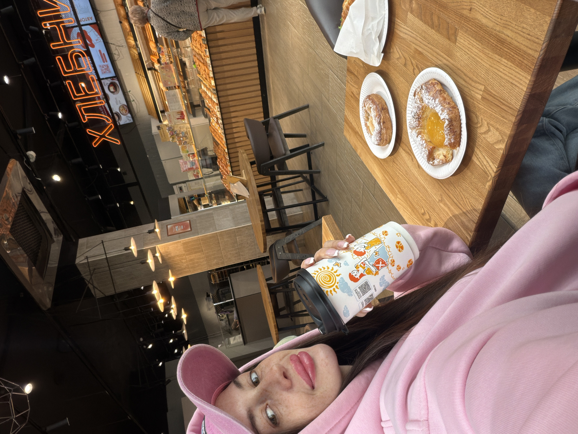 A woman in a pink hoodie and cap is holding a cup, likely in a bakery or cafe, possibly in Saint Petersburg, Russia. A wooden table displays two pastries on white plates and another pastry wrapped in white paper. In the background, customers browse a bakery display case illuminated by lights, with a neon sign reading "ХЛЕБНИШ." The interior features dark ceilings with modern lighting fixtures and wooden furnishings.